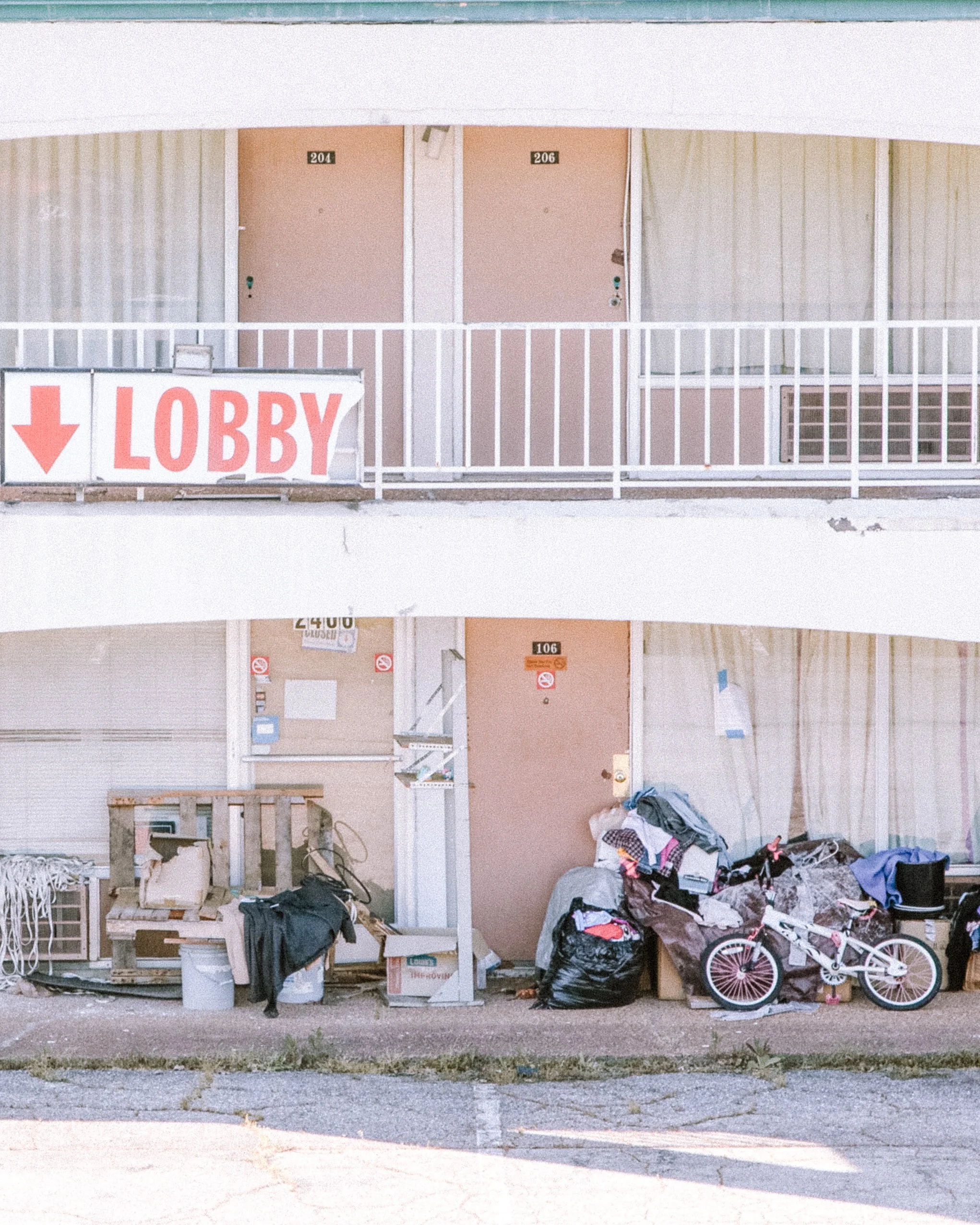 An apartment complex with two levels of doors, numbered 204 and 206, with a small balcony in front of each door. Several piles of garbage, bags, and miscellaneous items are on the ground in front of the building, including a pink and white children's