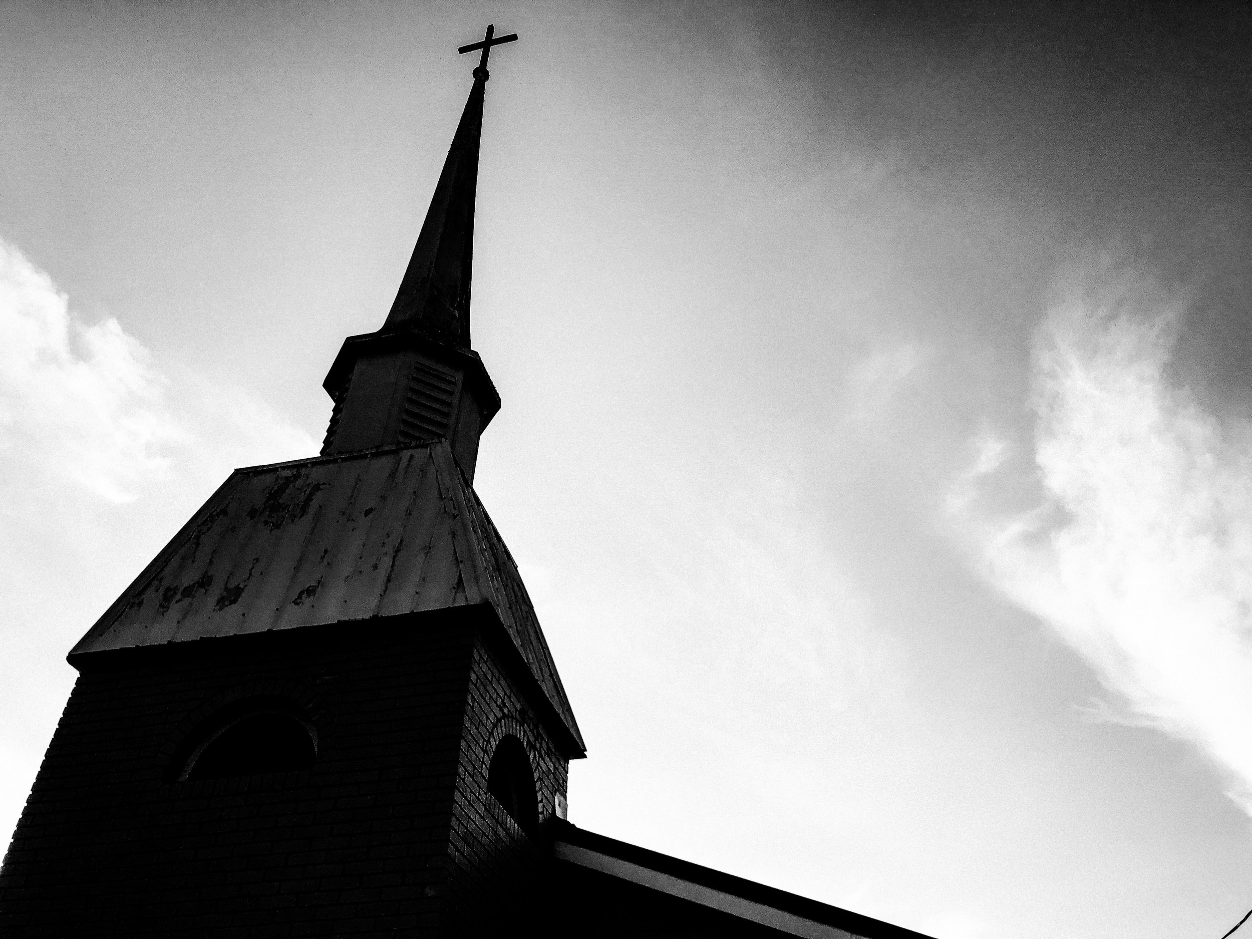 Black and white photograph of a church steeple with a cross on top, set against a cloudy sky.
