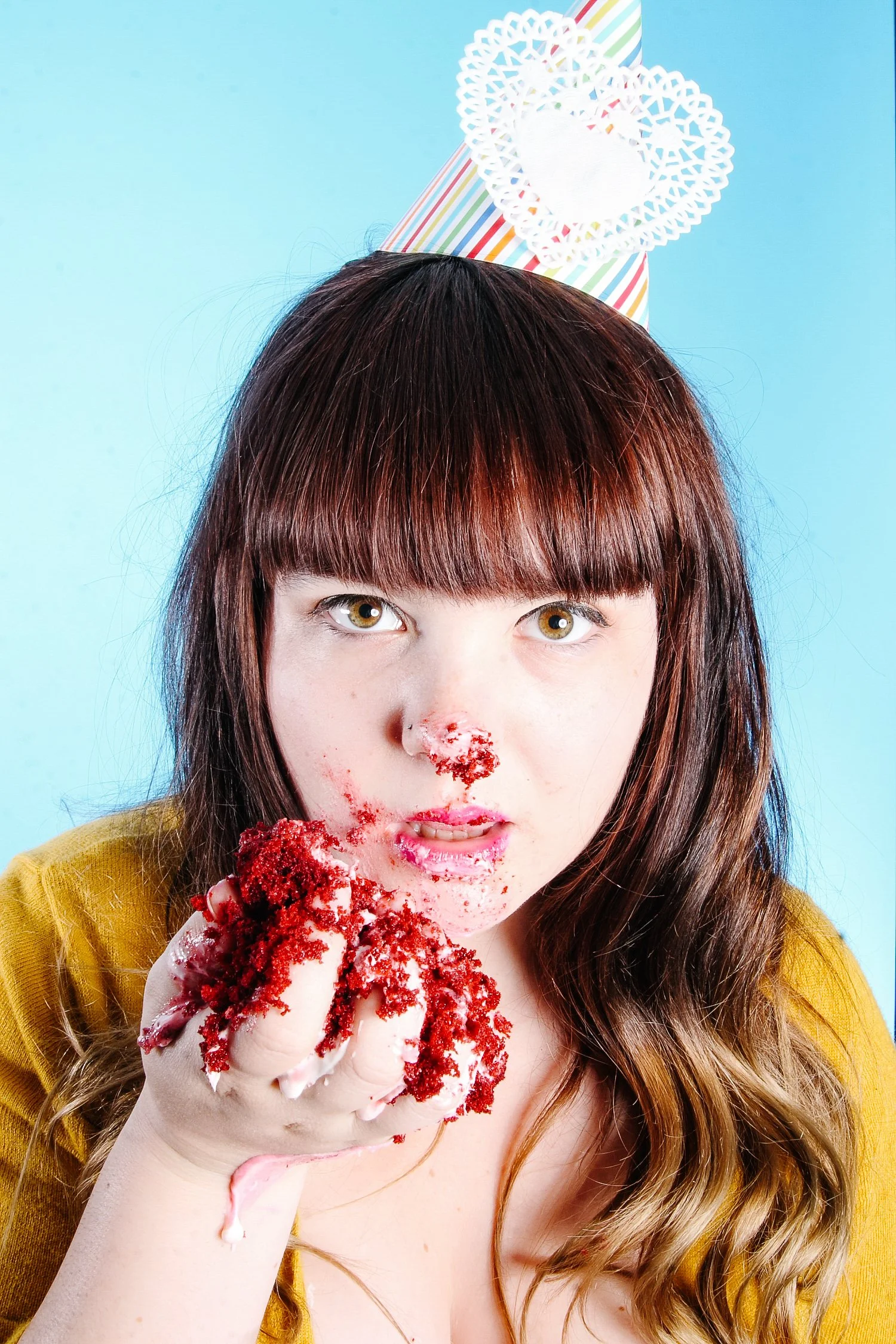 A young woman with brown hair wearing a yellow top is eating a cake with red crumbs and pink cream inside, with some crumbs on her face and hand, against a blue background with a colorful party hat above her head.
