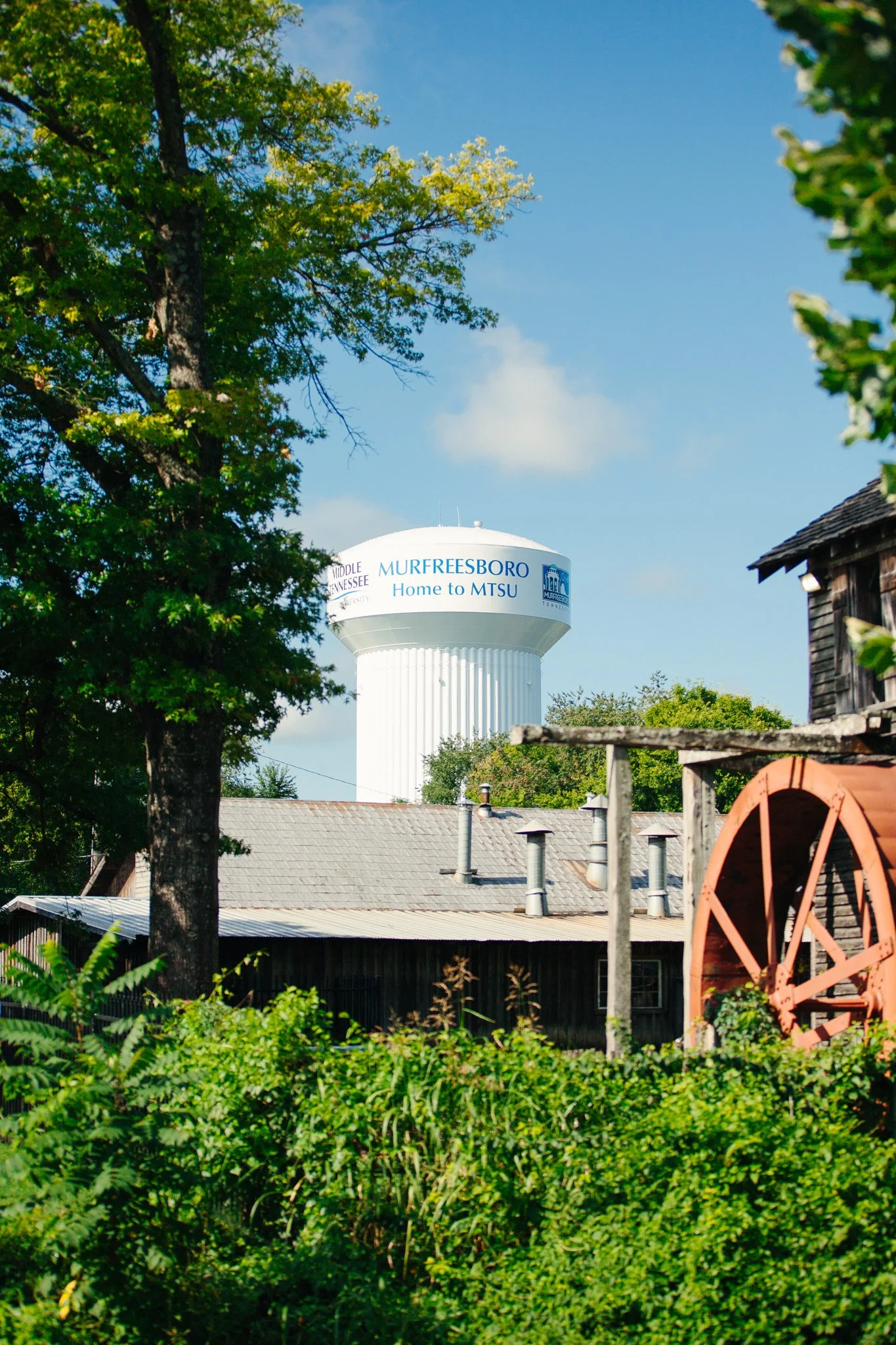 A water tower labeled 'Murfreesboro Home to MTSU' is seen above houses with gray roofs; trees with green leaves, a partially visible red windmill, and a wooden building are in the surroundings, under a clear blue sky.