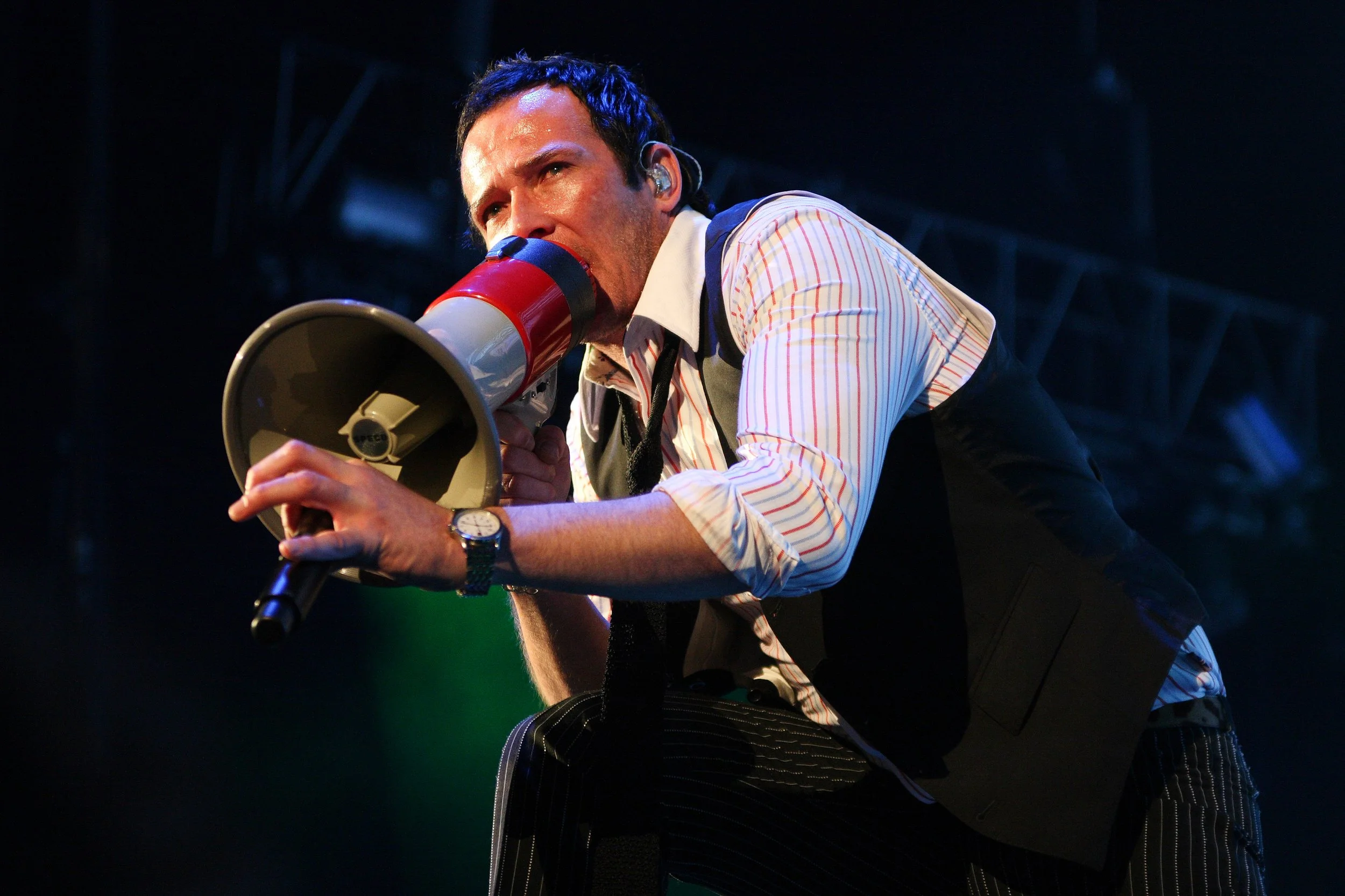 A man with dark hair and a beard is speaking into a red and white megaphone on a stage. He is wearing a white shirt with thin red and blue stripes and a black vest. The background is dark with some stage lighting.