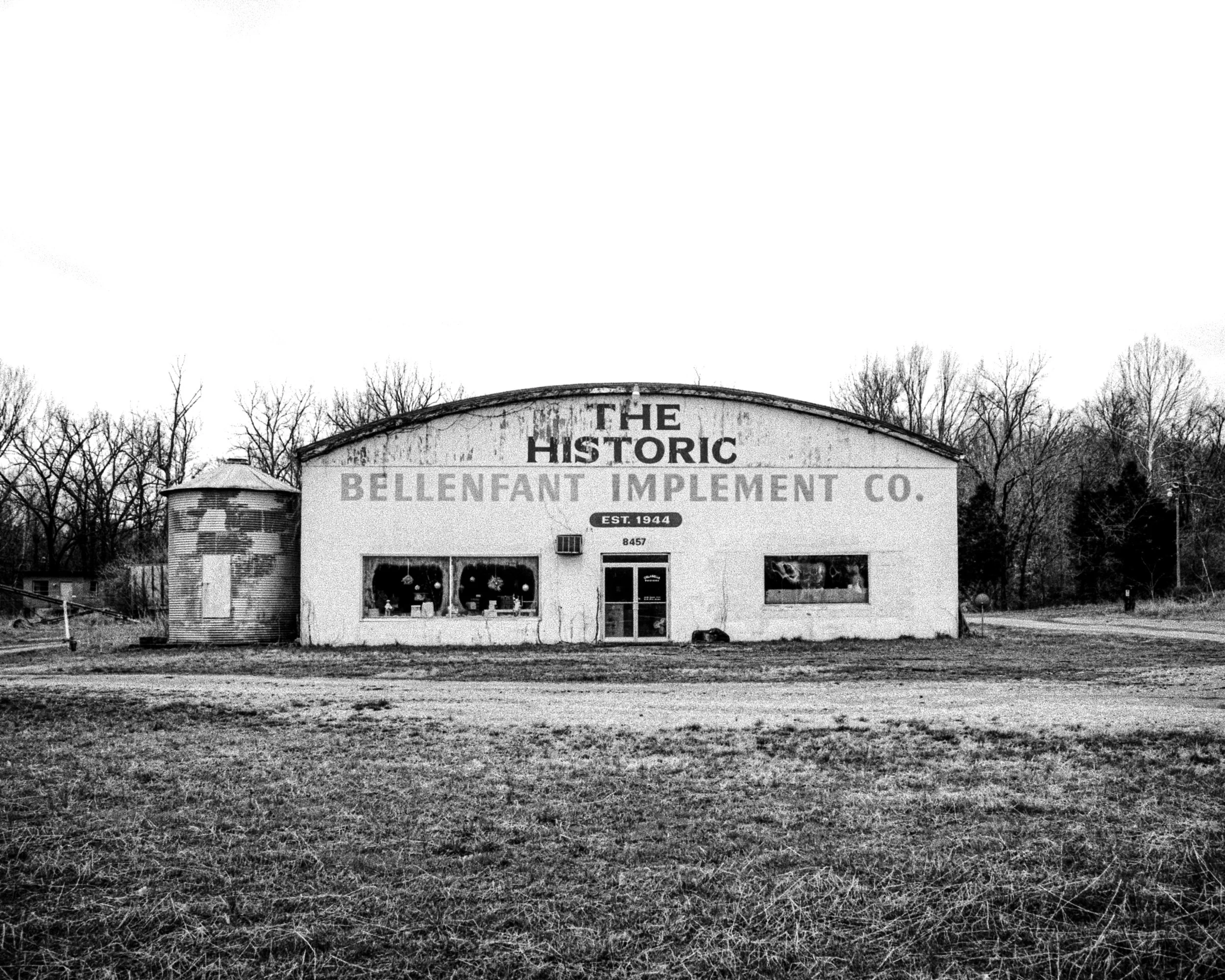 Black and white photo of a building with a sign reading "The Historic Bellenfant Implement Co." with trees behind it and a grassy open area in front.
