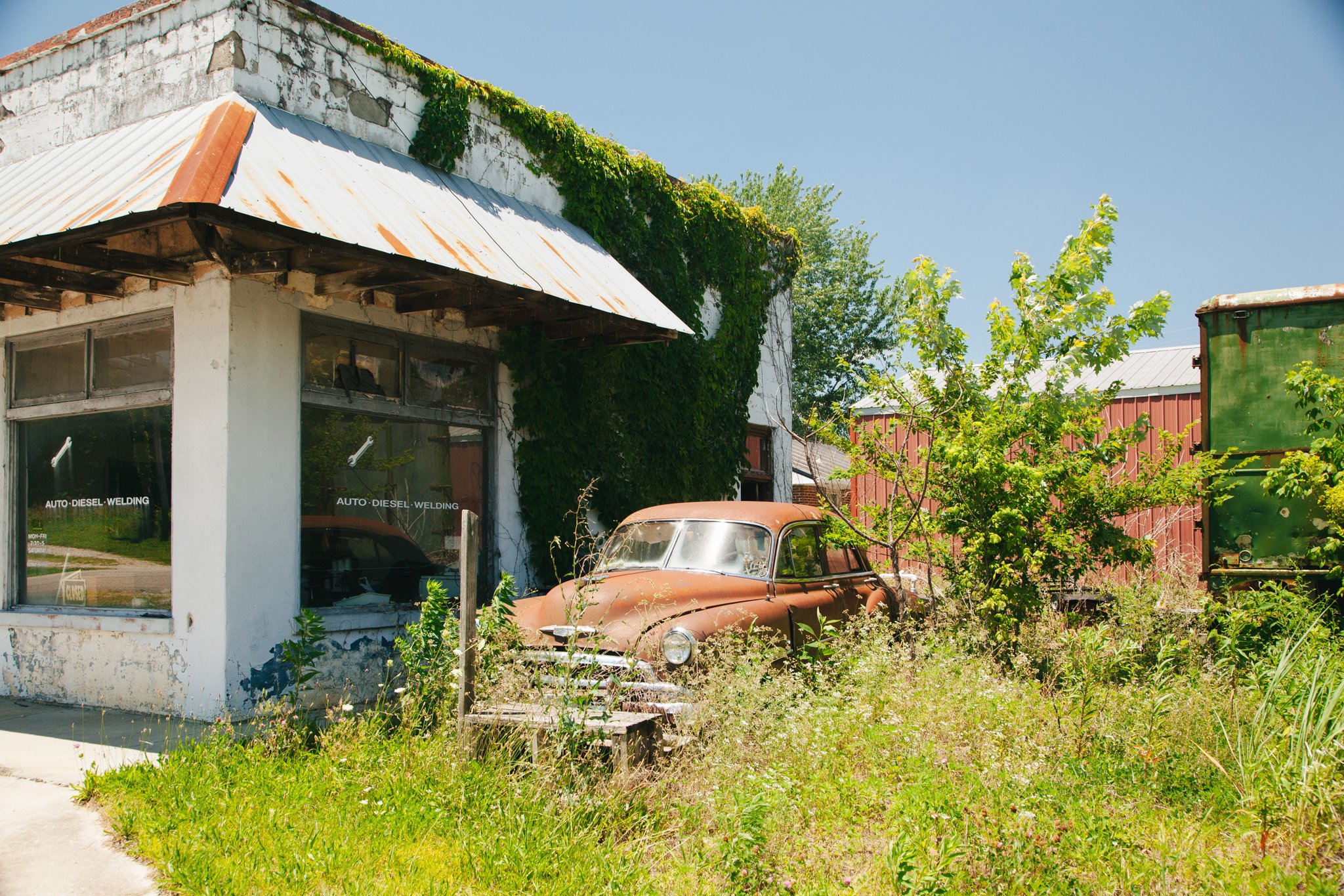 An old, rusty car parked in front of a weathered building with a partially broken window and overgrown plants, including ivy and small trees, with a clear blue sky in the background.