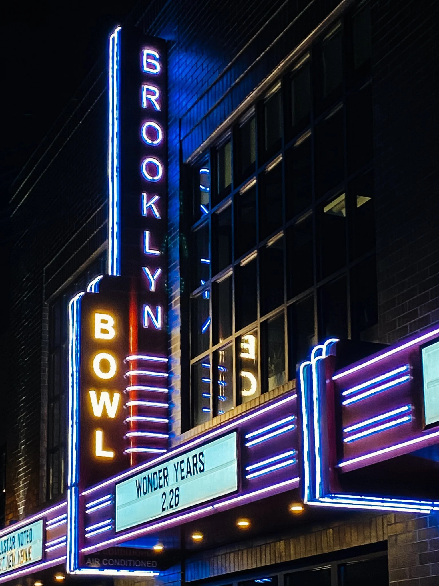 Nighttime view of a movie theater marquee with neon lights and sign reading 'Brooklyn' and 'The Wonder Years' showing on the digital display.