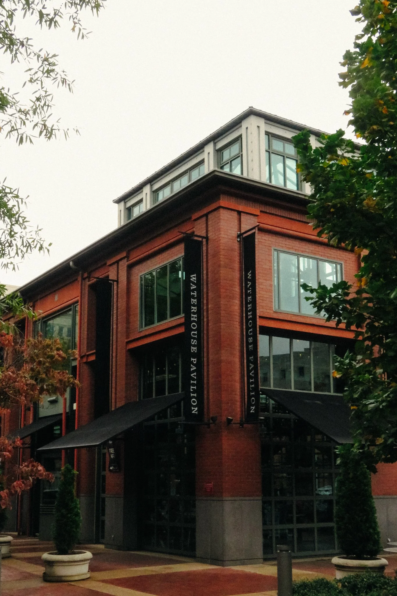 A multi-story building with a brick exterior, large windows, and hanging banners that read 'Waterhouse Pavilion.' The building is surrounded by trees and potted plants, with a sidewalk and outdoor seating area in front.