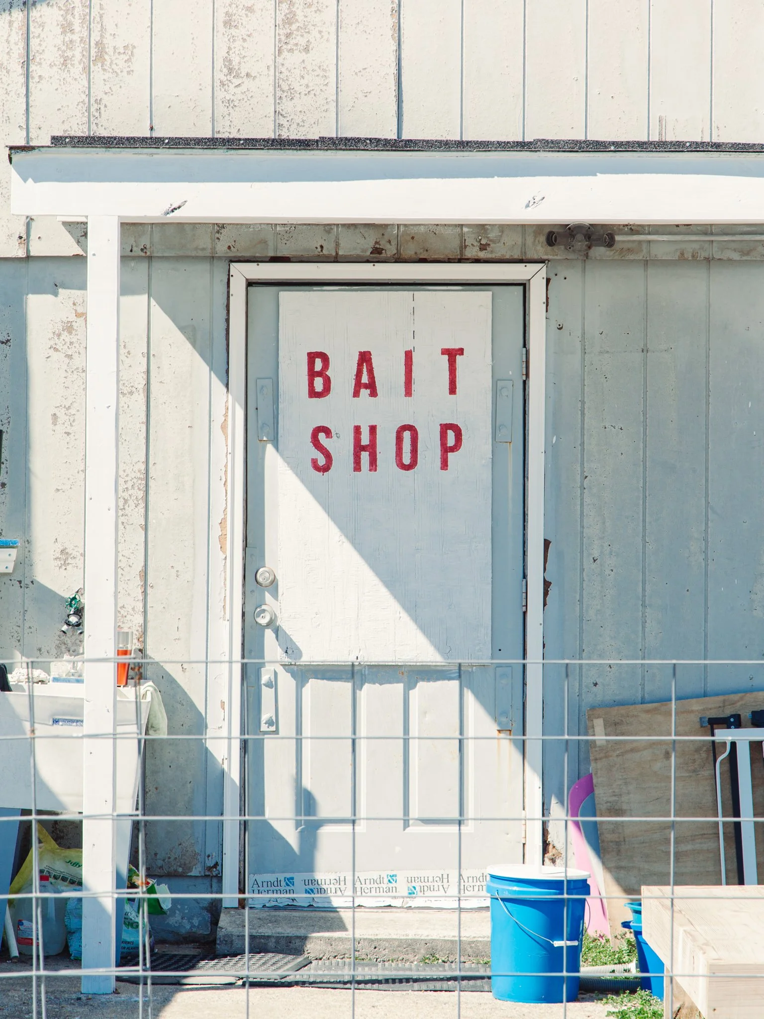 A white door with red lettering that says 'BAIT SHOP,' surrounded by construction materials and a chain-link fence, with a blue bucket and pink object in front.