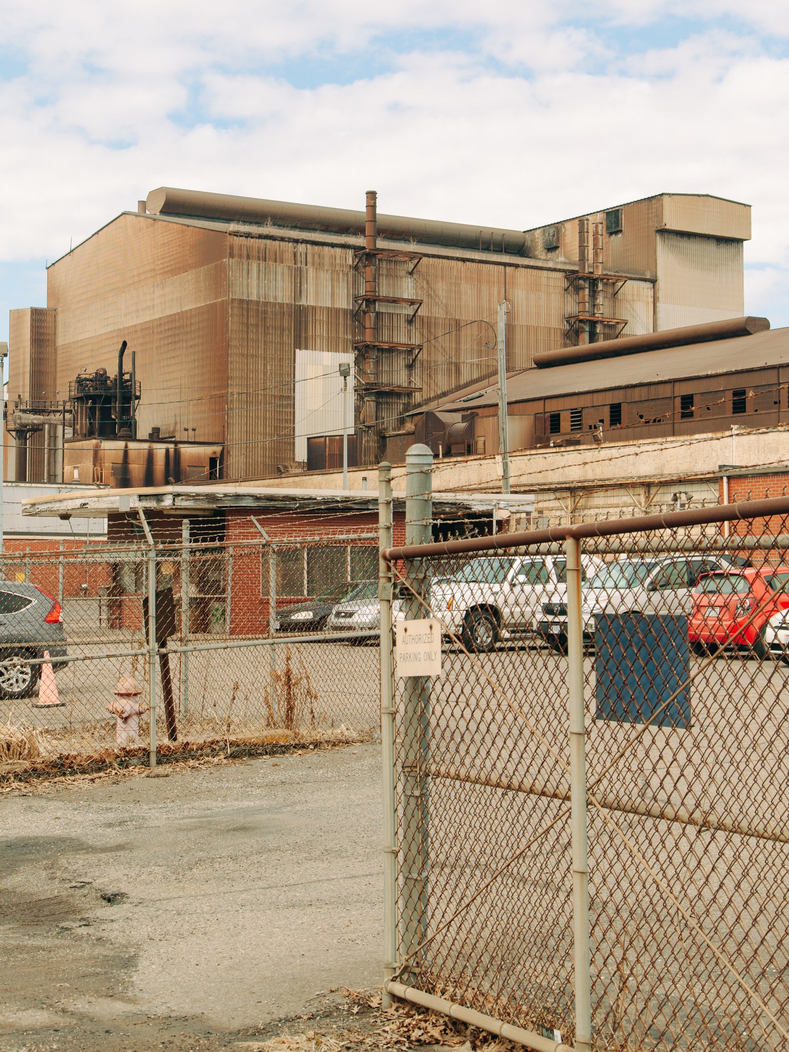 An industrial building with rusty metal walls surrounded by a chain-link fence, parked cars, and a sign that says 'Authorized Parking Only'.