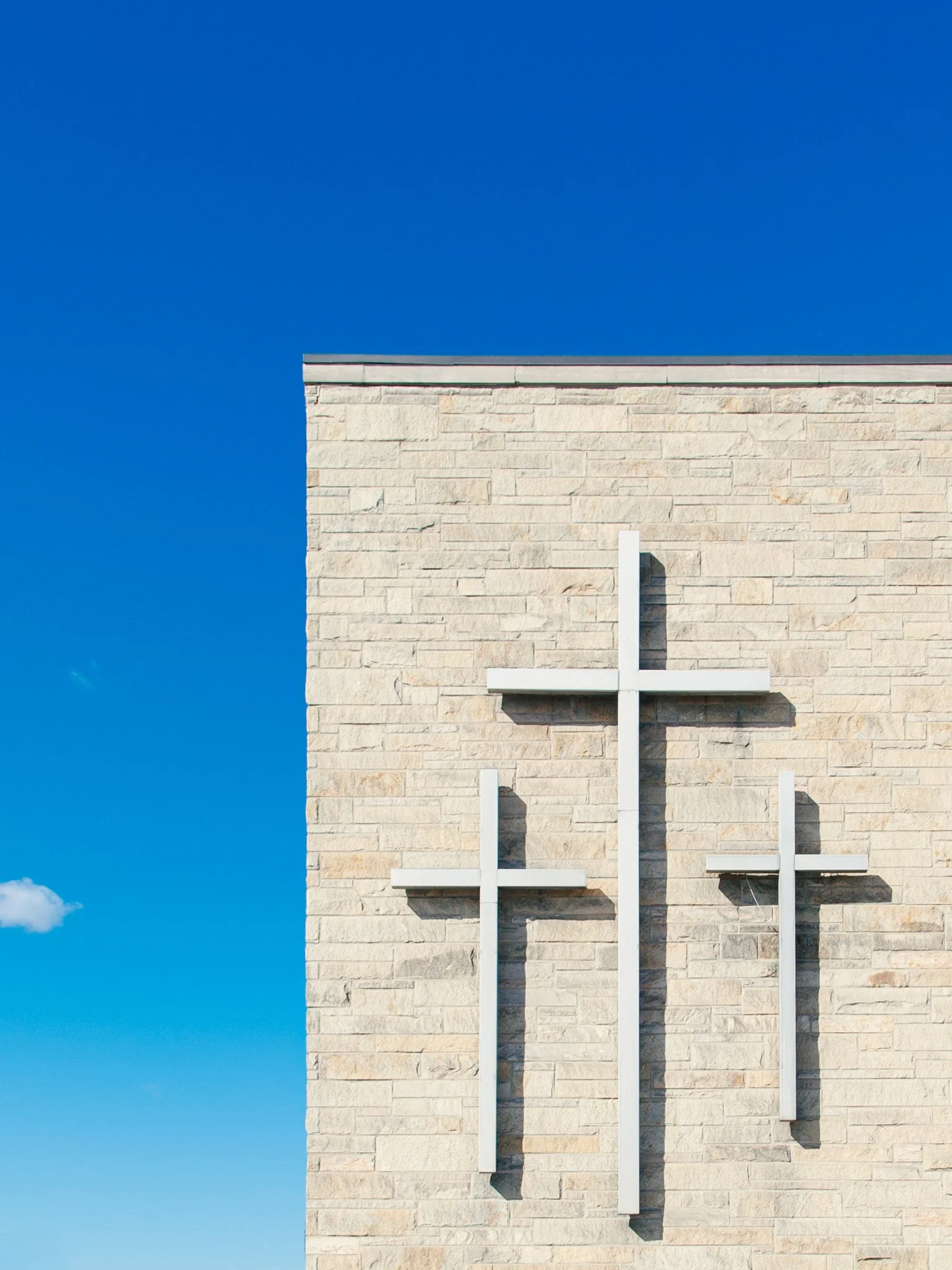 The front of a beige brick church building with three white crosses mounted on the wall and a blue sky in the background.