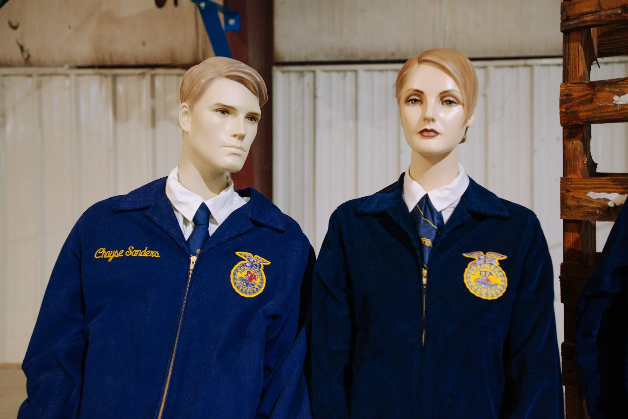 Two mannequins dressed in blue FFA jackets. The left mannequin has the name tag 'Craye Sanders' and the right one has a similar FFA emblem on their jacket. They are positioned indoors with a beige wall and wooden structure in the background.