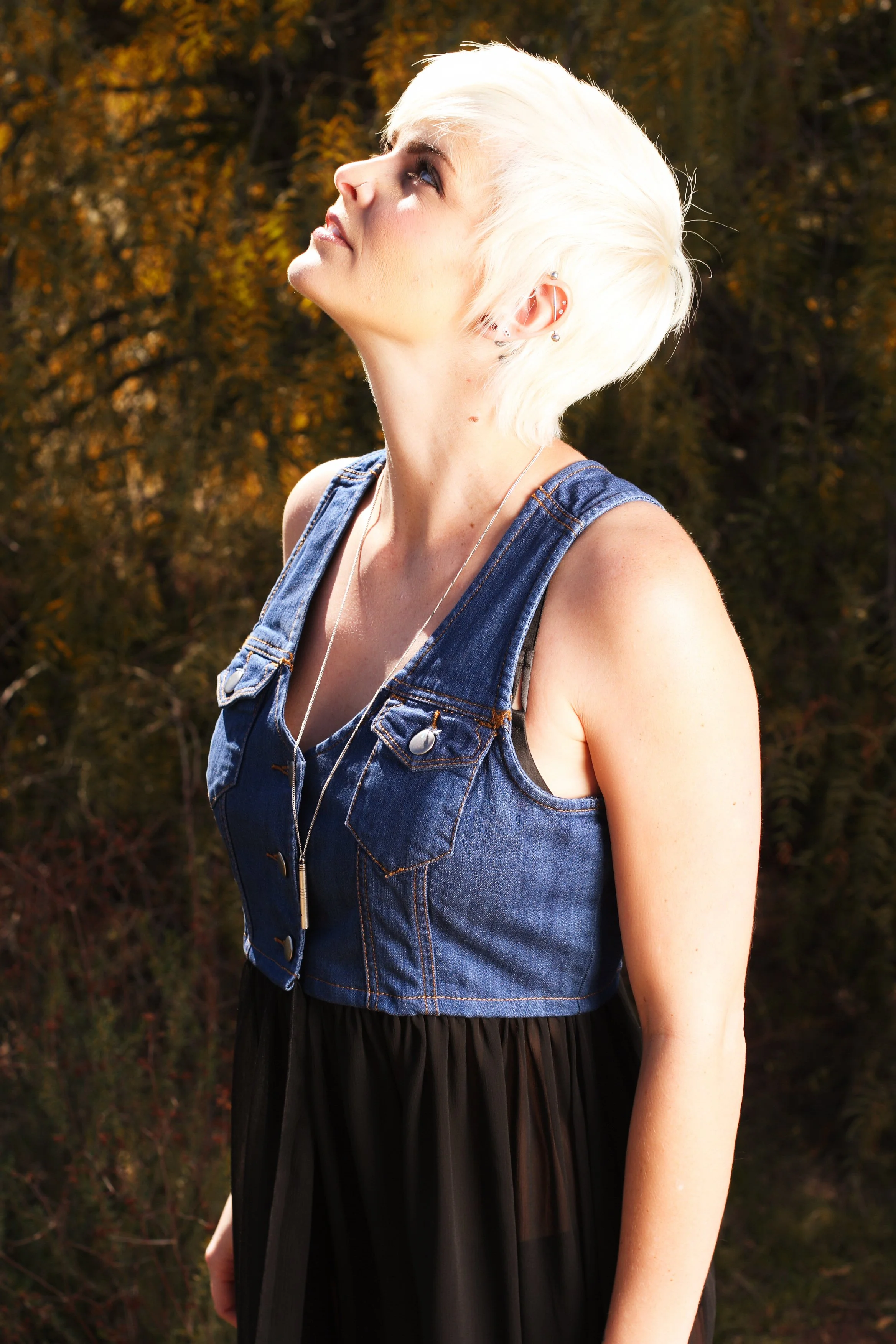 A woman with short platinum blonde hair, wearing a sleeveless denim jacket over a black dress, standing outdoors with autumn-colored trees in the background. She is looking upward with her face slightly tilted.