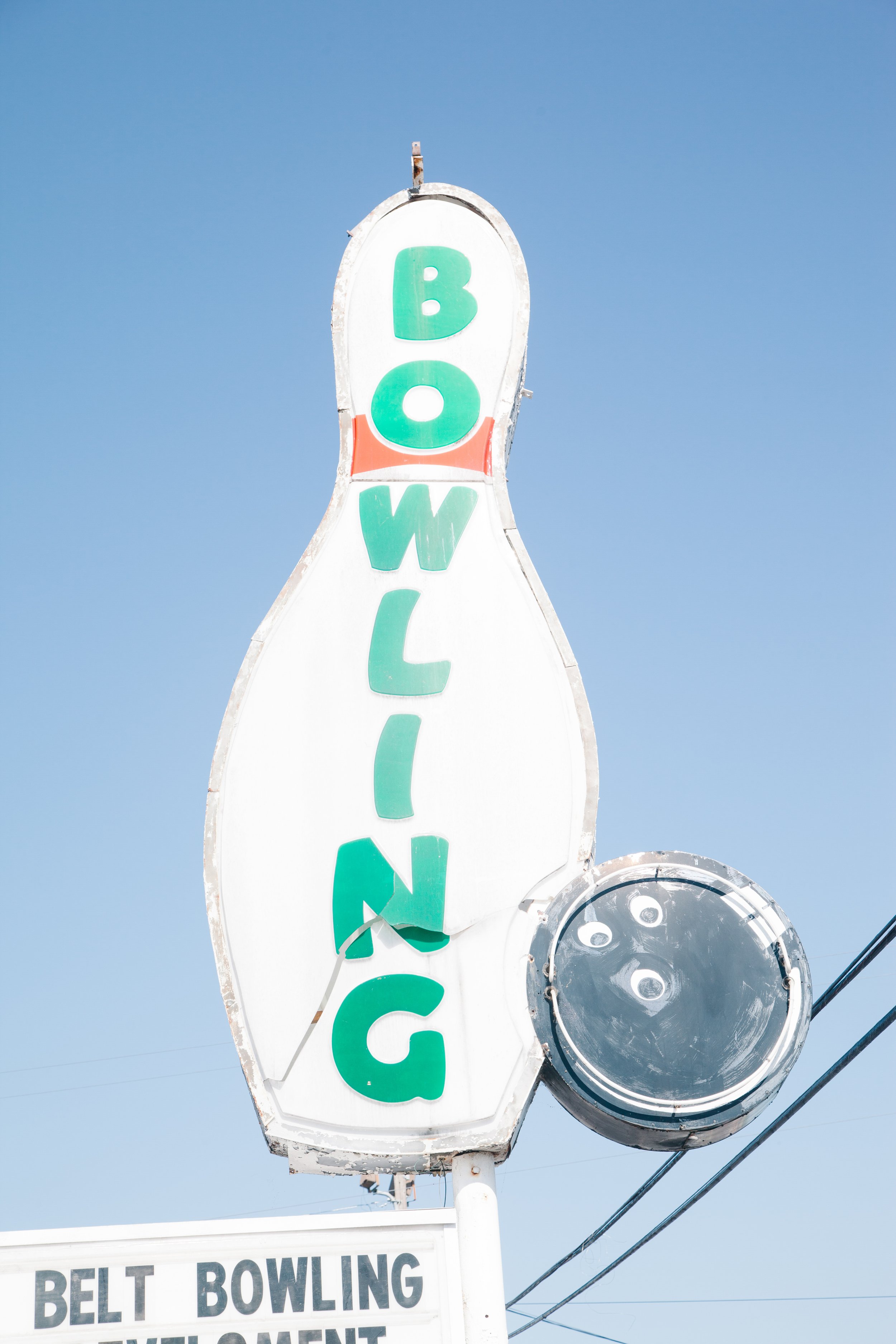 Vertical sign with the word "BOWLING" in green letters, a circular light fixture at the bottom, and a billboard in the background reading "BELT BOWLING." Clear blue sky.