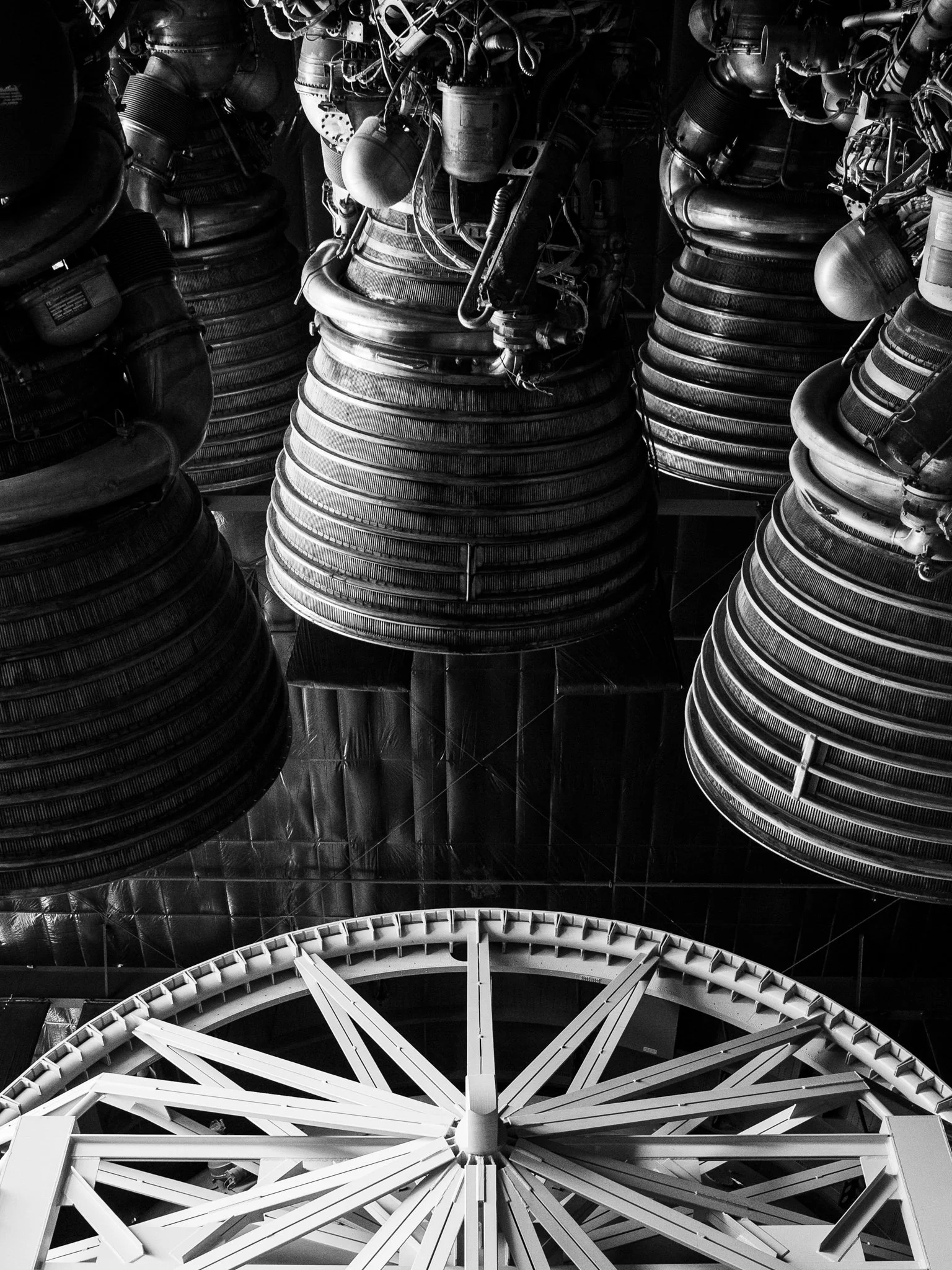 A black and white photo of a large Ferris wheel, viewed from below, with its white spokes and central hub prominently visible. The background shows an industrial setting with large machinery and pipes.