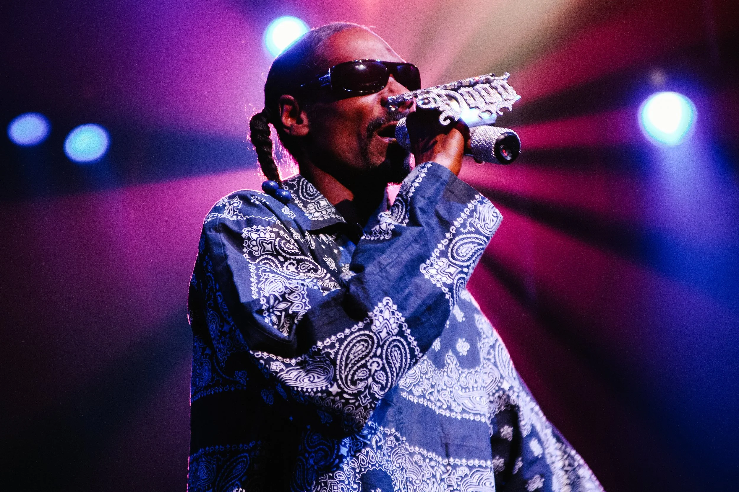 A performer on stage wearing dark sunglasses, a patterned shirt, and holding a glittery microphone. The background features colorful stage lighting.