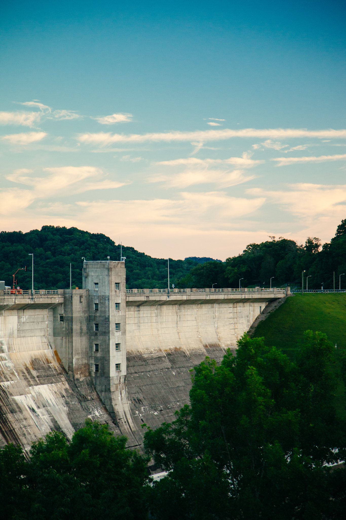 A large concrete dam with a curved face, trees in the foreground, a hill covered with trees in the background, and a partly cloudy sky.