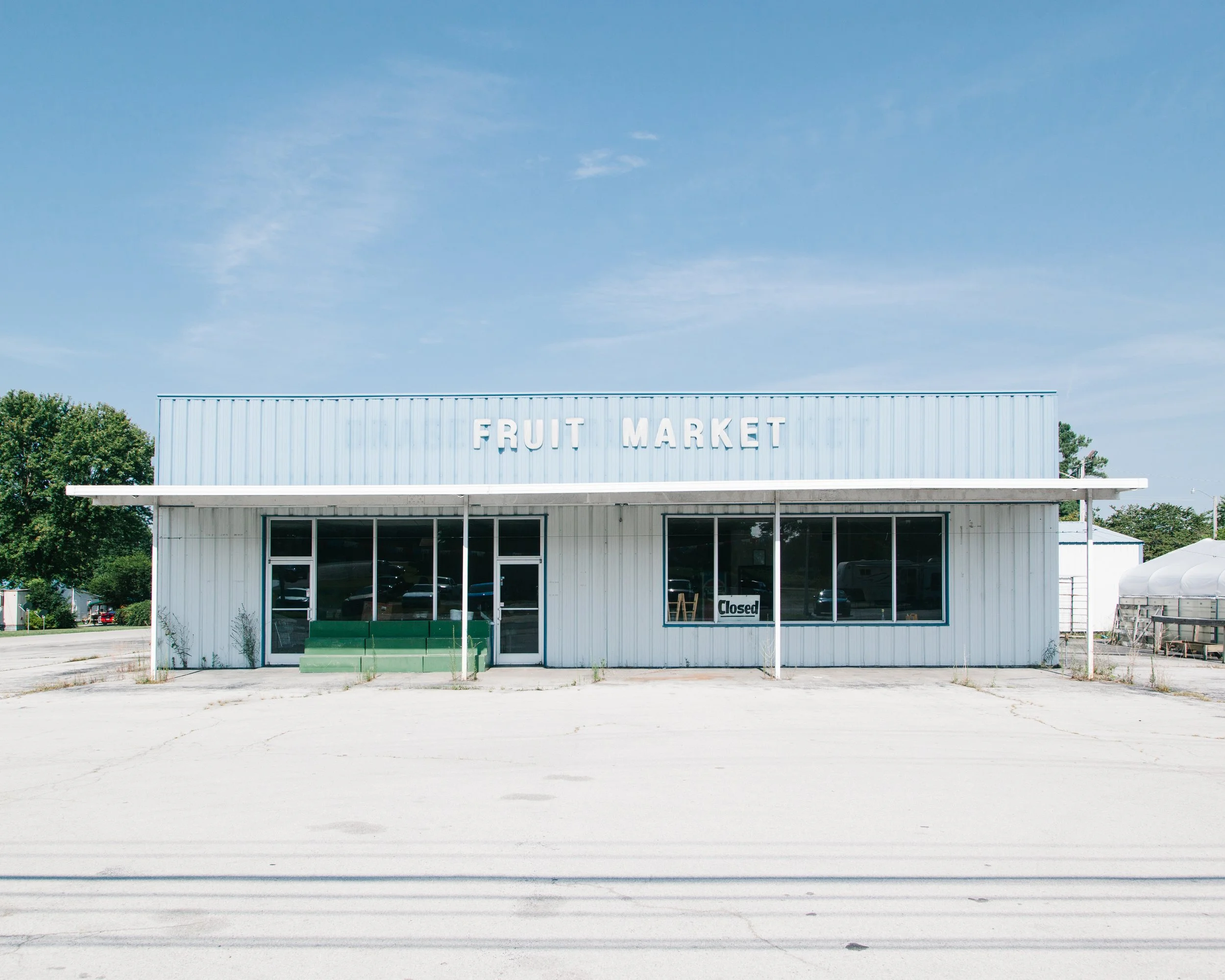 Empty storefront with a blue metal facade reading 'Fruit Market' in white letters, large glass windows and door, small green bushes in front, and a "Closed" sign on the door under a partly cloudy sky.
