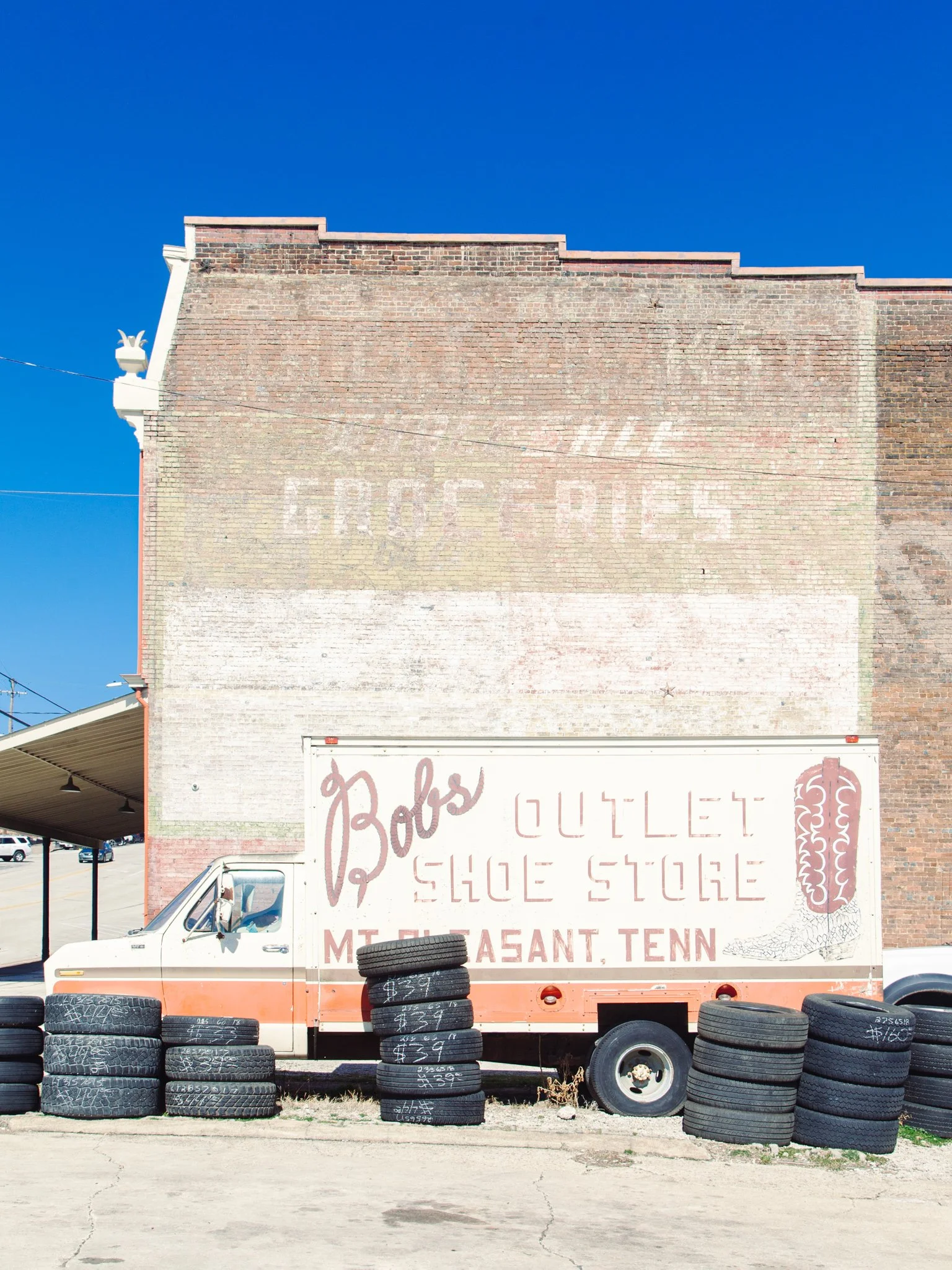 White and orange delivery truck with tires stacked in front, parked in front of a large faded brick wall advertising Bob's Outlet Shoe Store in Mount Pleasant, Tennessee, with a clear blue sky overhead.
