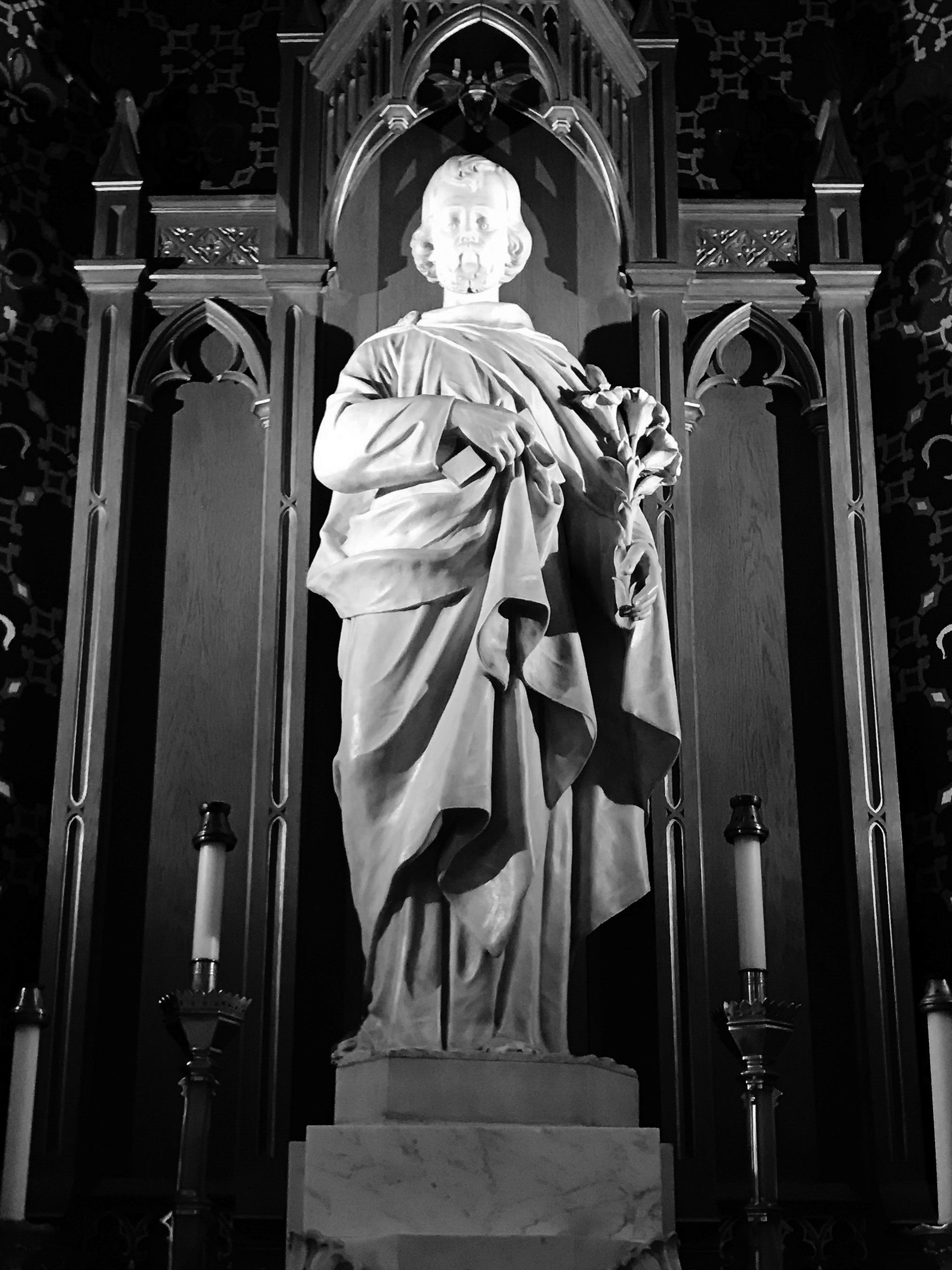 A black and white photo of a statue of Jesus Christ holding a bouquet of lilies, standing on a pedestal in a church, with ornate Gothic-style architecture behind.