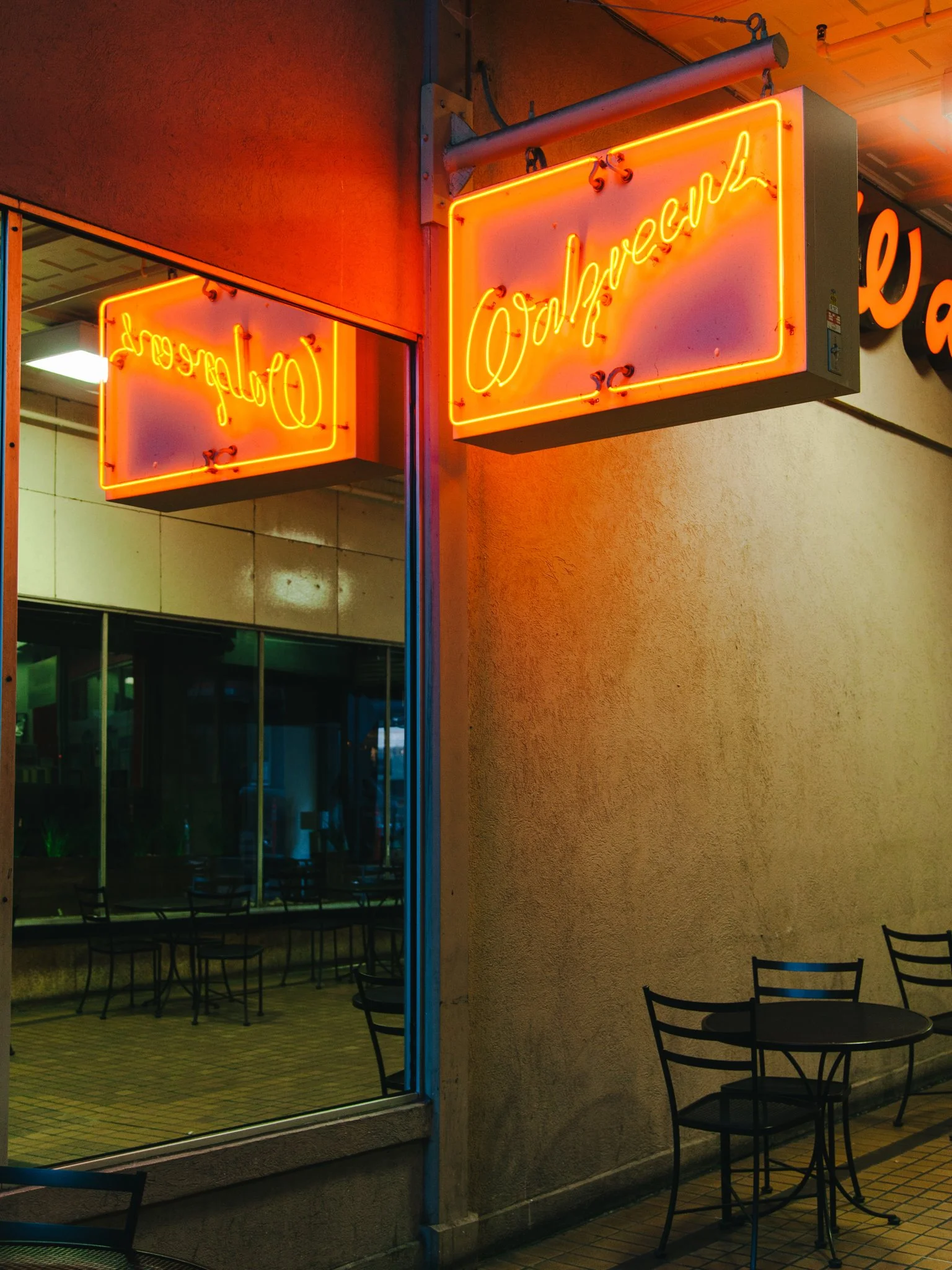 Neon sign in orange says 'Open' and is mounted on a wall outside a restaurant or cafe with patio seating. The patio has black metal chairs and tables, and indoor lighting is visible through the large window.