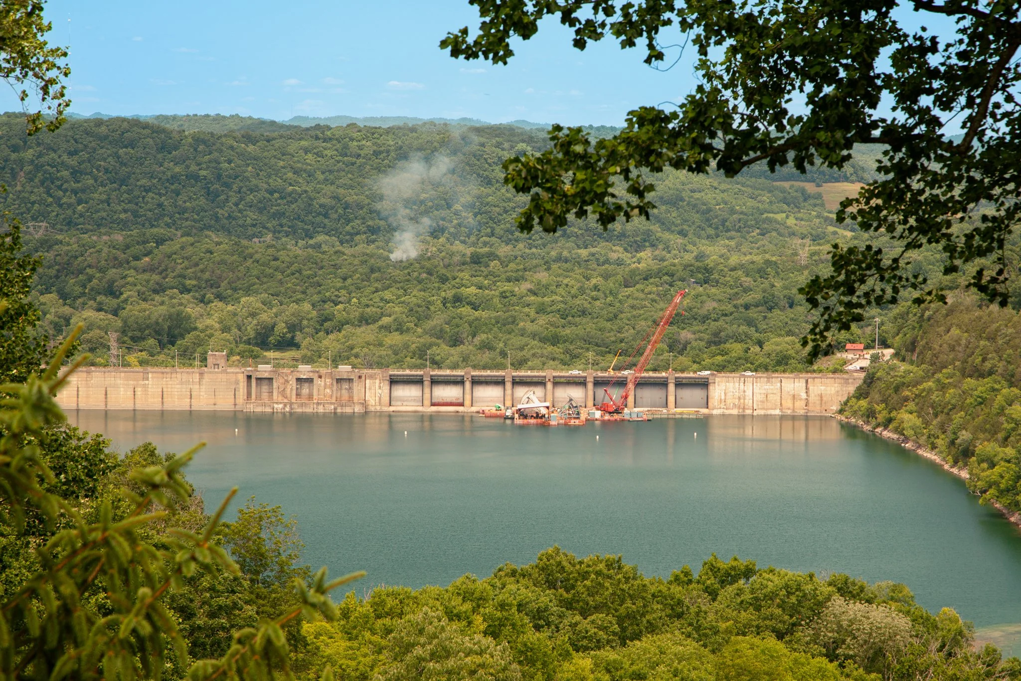 A large dam with water reservoir in front of a green forested hillside, with construction cranes and machinery at the dam site.