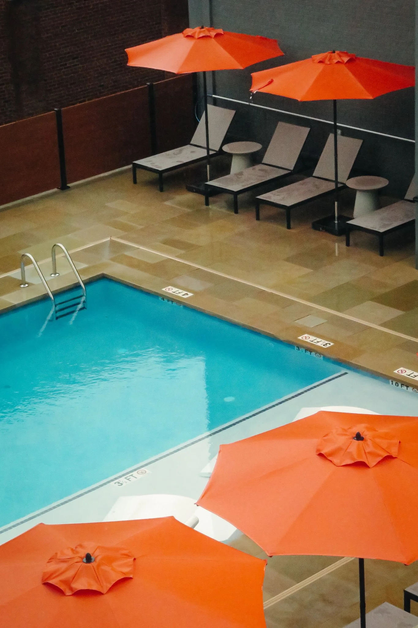 Poolside lounge chairs with orange umbrellas and a swimming pool with a ladder in an indoor setting.