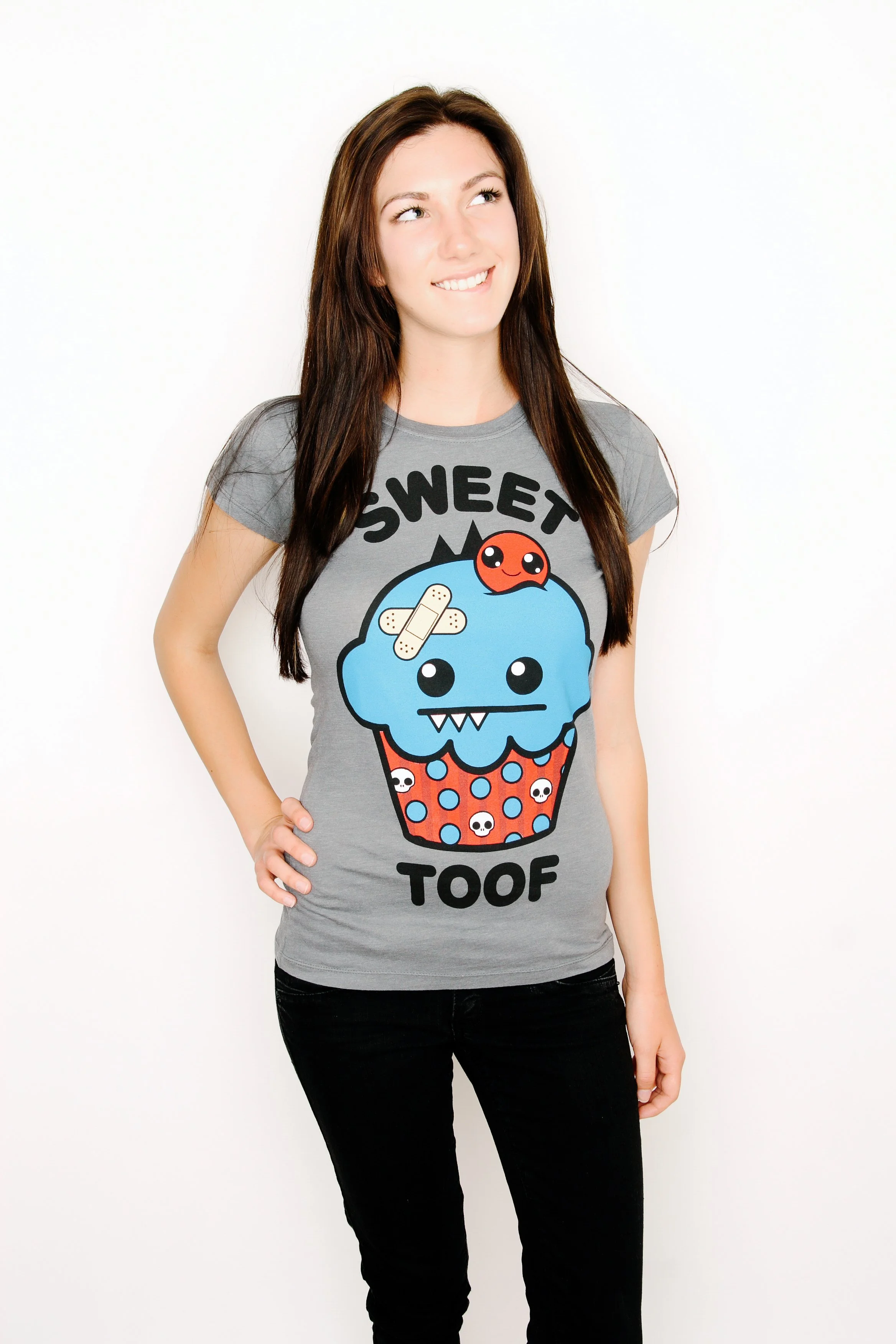 A woman with brown hair wearing a gray T-shirt with a cartoon cupcake design and the words 'SWEET TOOF'. She is standing against a white background and smiling while looking to her right.