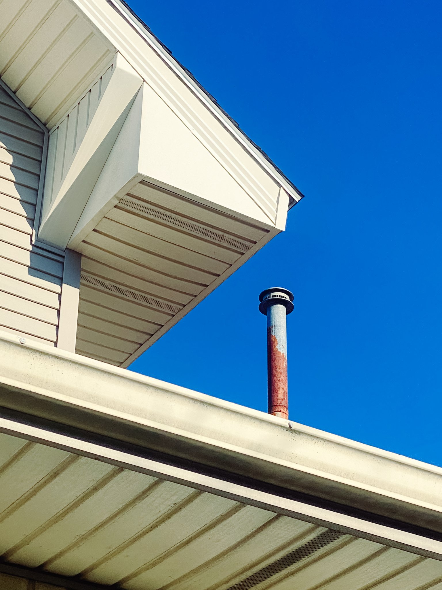 Close-up of the corner of a house with beige vinyl siding and white trim, showing part of the roof, a rusty red chimney pipe, and a clear blue sky in the background.