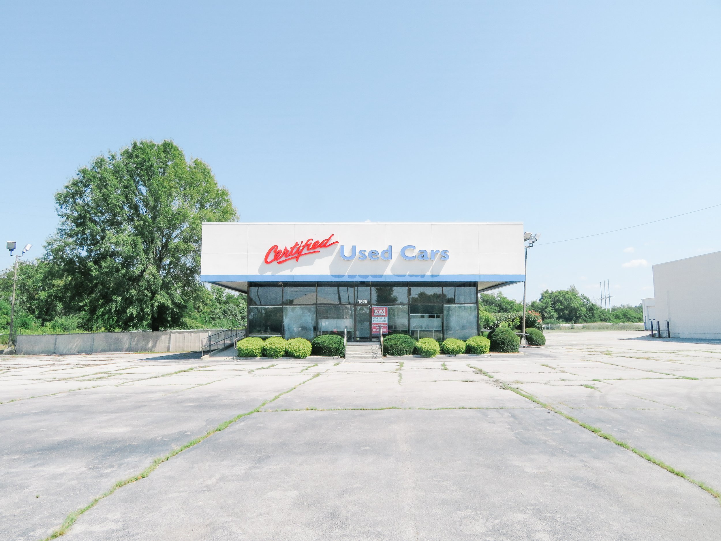 Front view of a car dealership with a sign that reads 'Certified Used Cars', a clear sky, green trees, and a mostly empty parking lot.