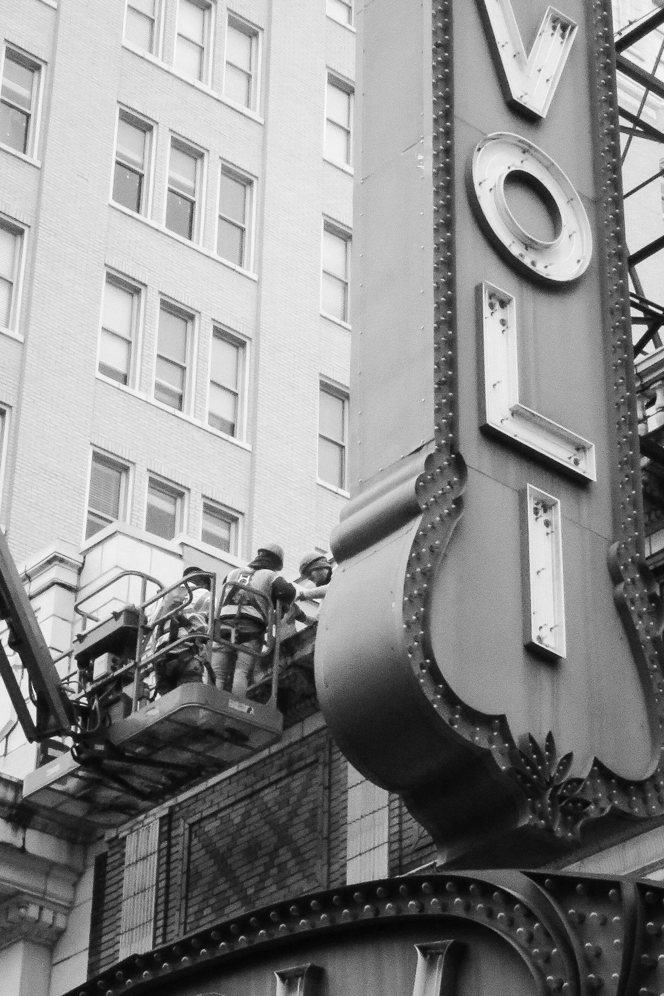 A close-up black and white photo of workers on a cherry picker repairing or maintaining a vintage-style marquee sign on a building in an urban area.