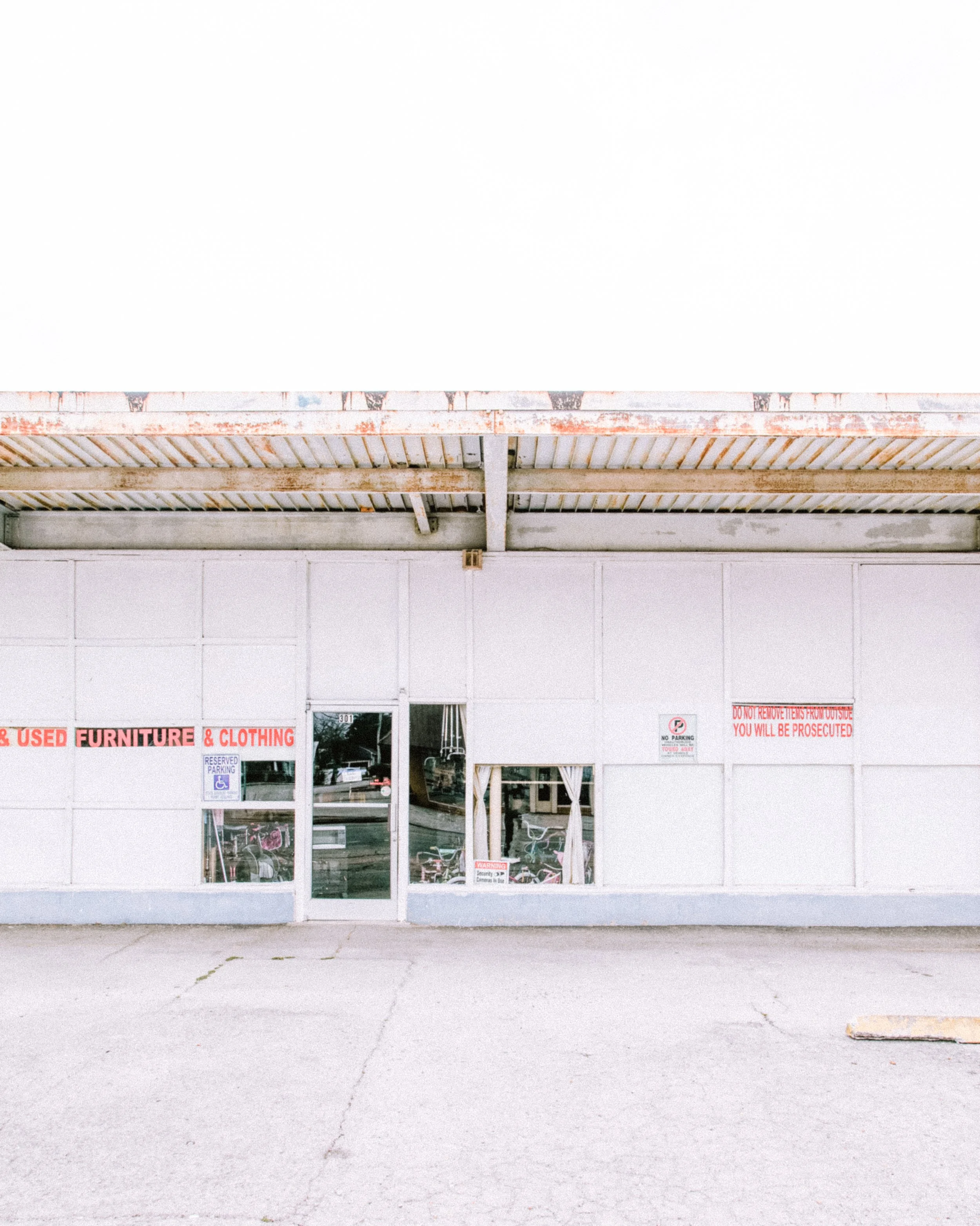 Front view of a thrift store with glass doors and signs that read 'USED FURNITURE & CLOTHING' and other notices, situated in a commercial area with an empty parking lot.