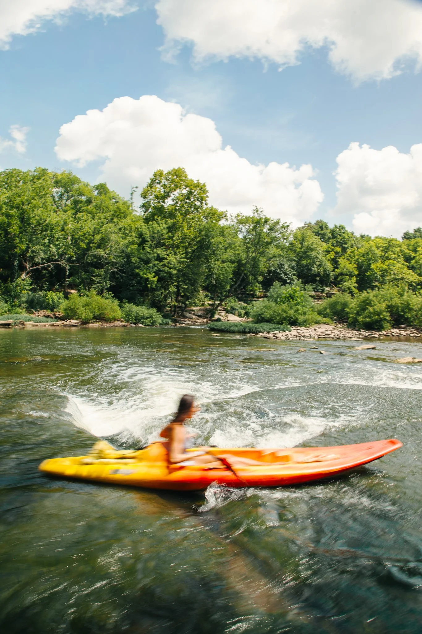 Person kayaking on a river surrounded by green trees under a partly cloudy blue sky.