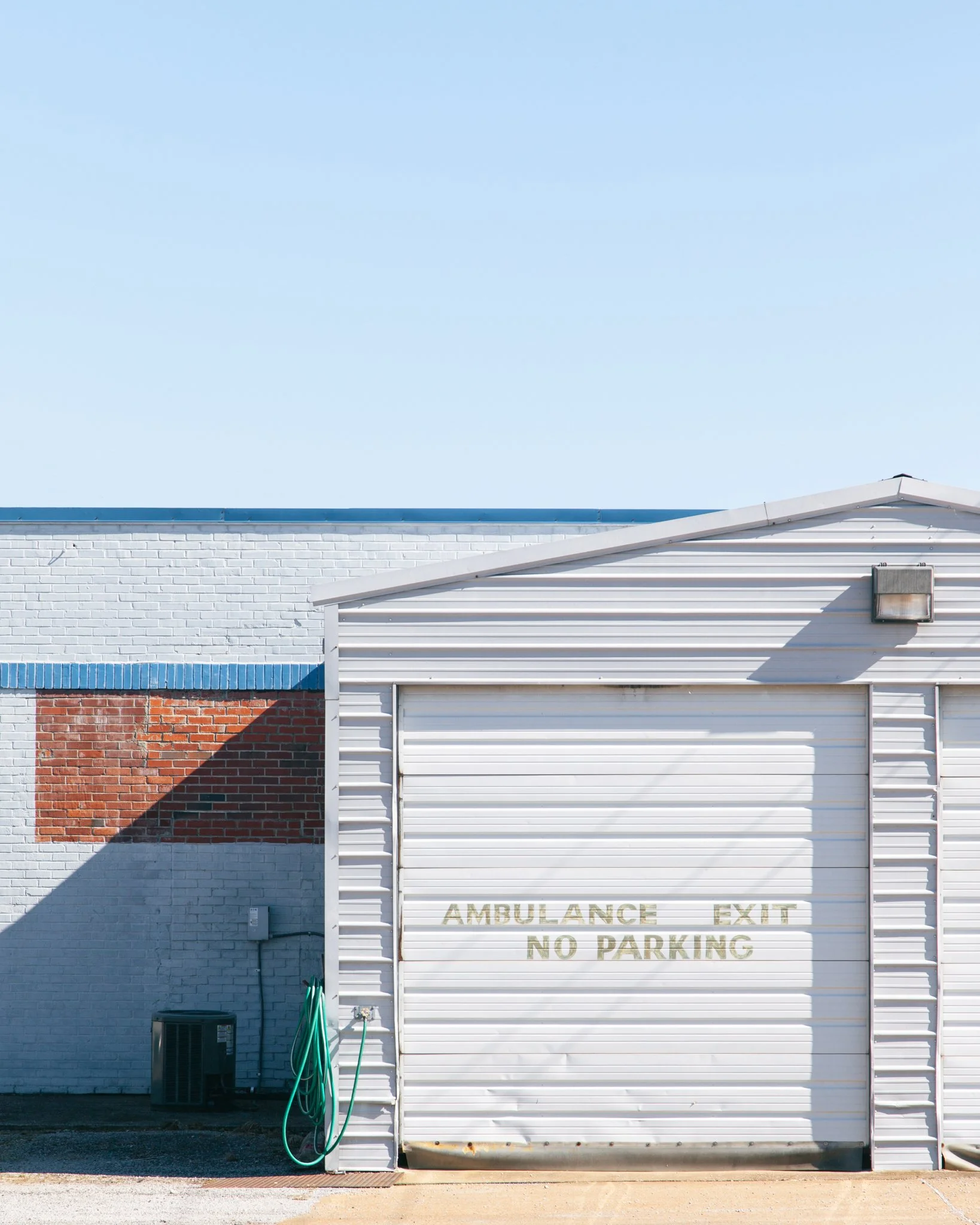 White building with a roller shutter door labeled 'AMBULANCE EXIT NO PARKING.' The building has a brick section and a blue roof edge. Two green hoses are hanging on a wall, with a small black utility box at the lower left. The sky is clear and blue.
