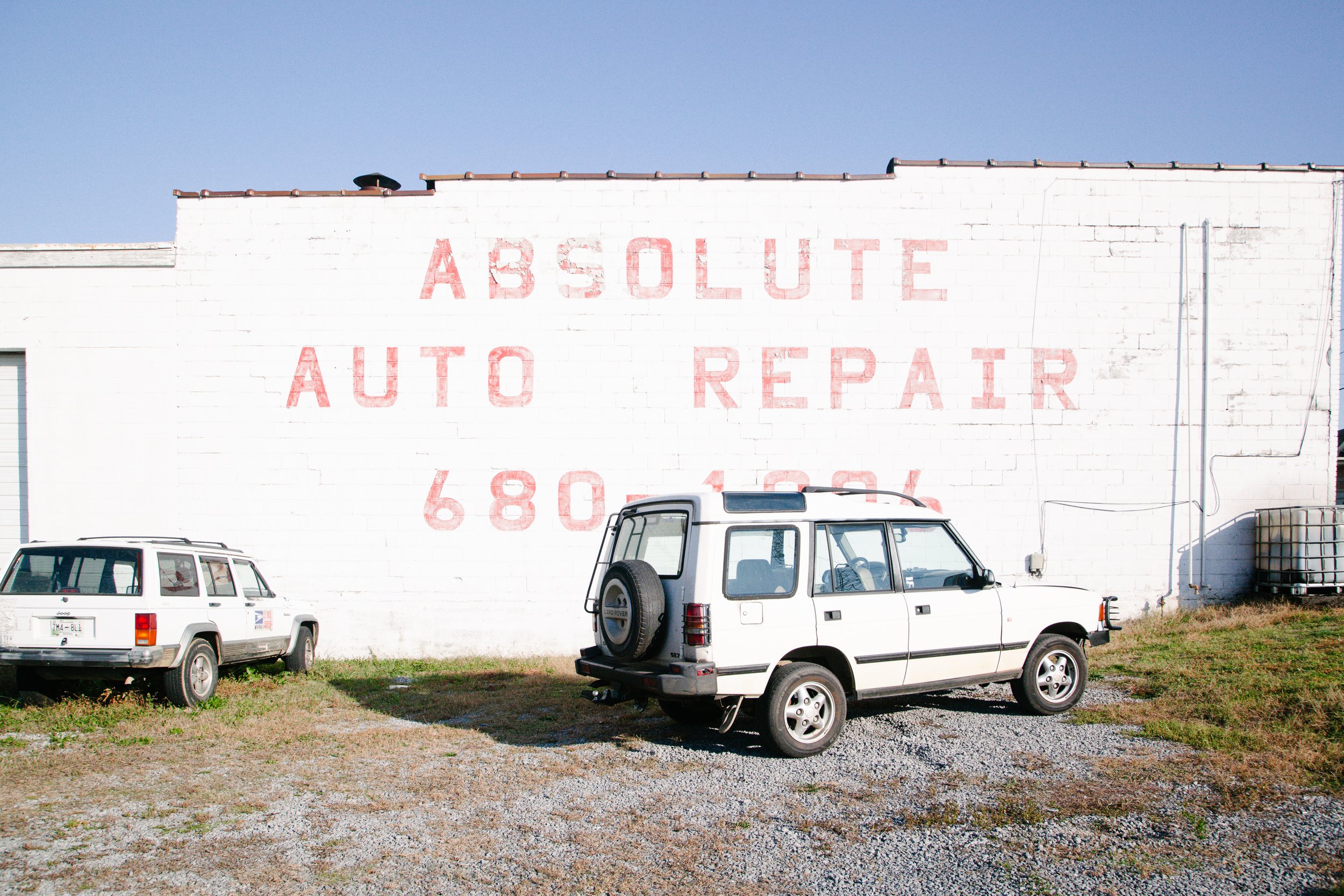 White brick building with red faded letters saying 'ABSOLUTE AUTO REPAIR 680-4604' on the wall, and three parked white vehicles in front on a gravel lot under a clear blue sky.