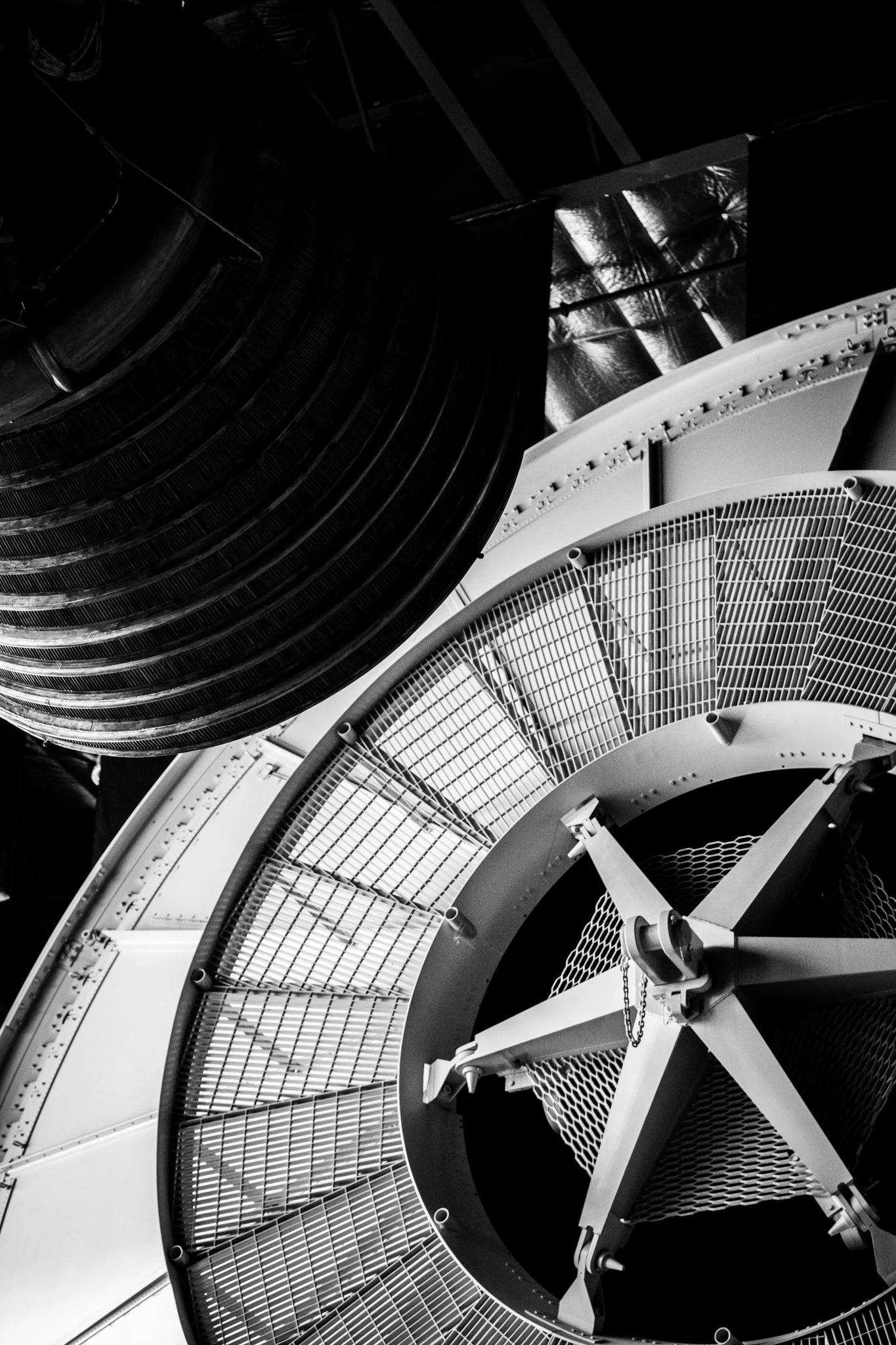 Close-up of a large industrial or scientific apparatus with circular metal grates, pipes, chains, and structural components in black and white.