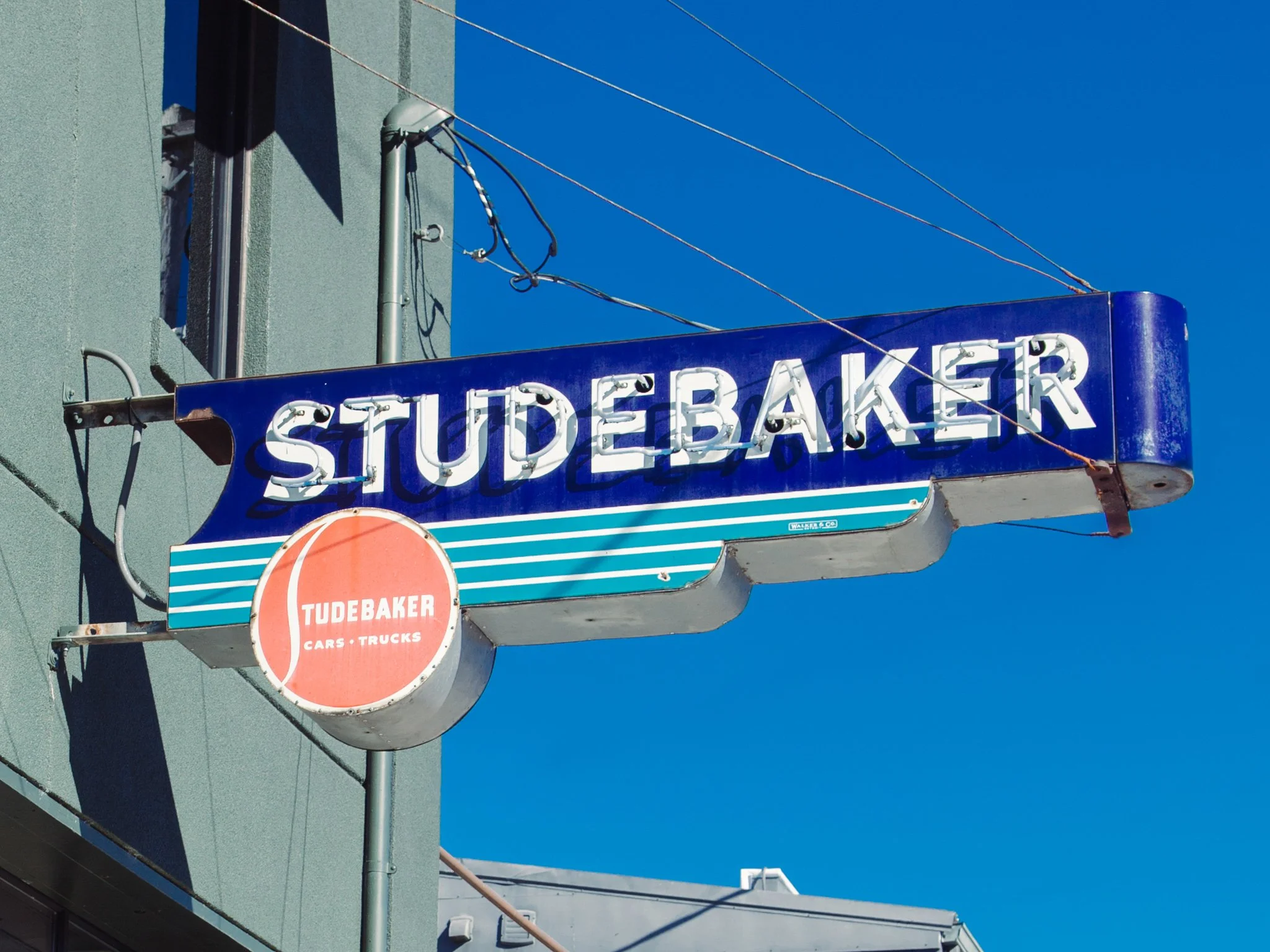 A blue and white neon sign reads 'STUDEBAKER' with a red circle below and the words 'TUDebaker CARS TRUCKS' inside. The sign is attached to the side of a building under a clear blue sky, with overhead wires and part of the building visible.
