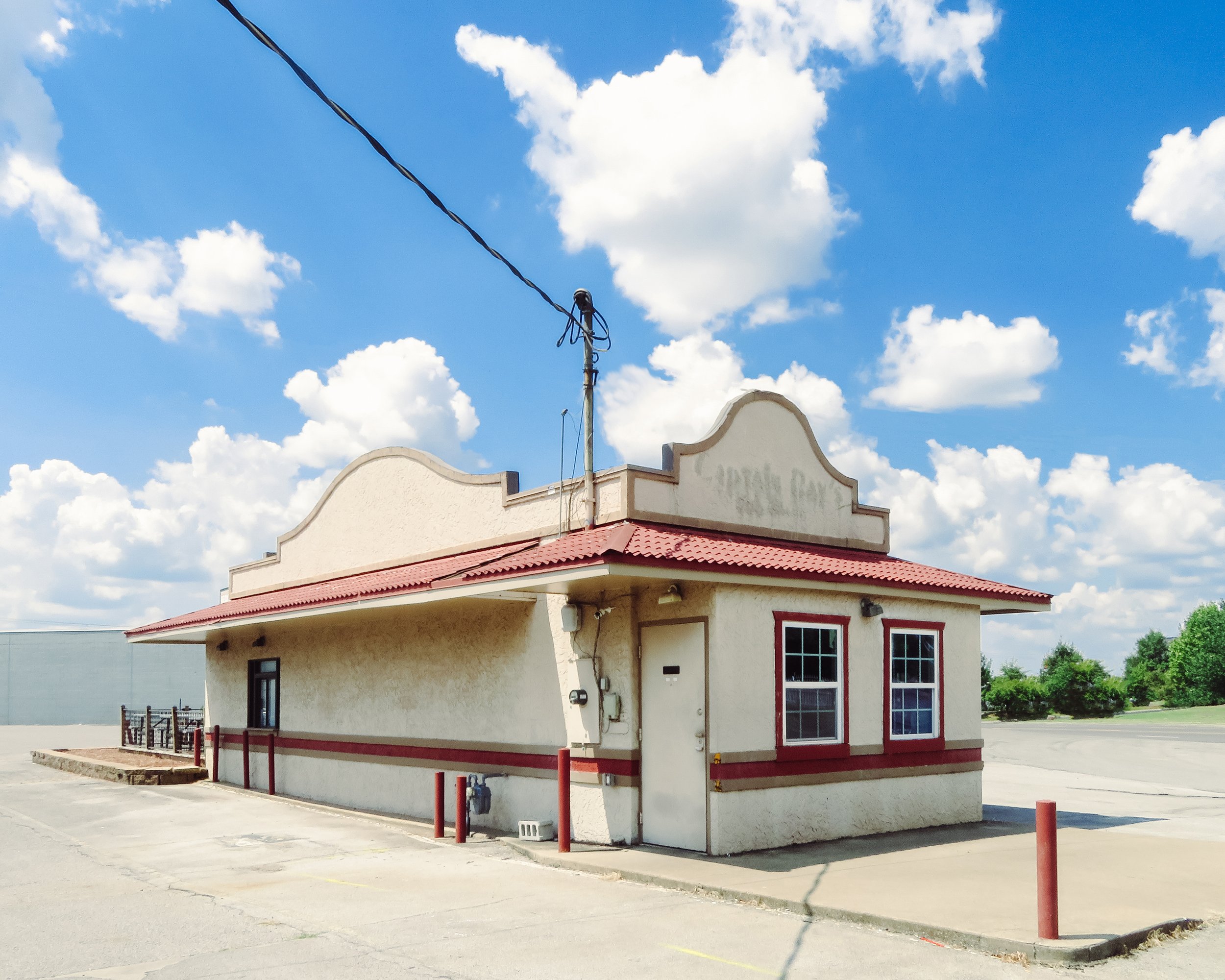 A single-story building with a red-tiled roof and white stucco walls, featuring red trim around the door and windows, situated in a parking lot with red bollards and a clear blue sky with scattered white clouds overhead.