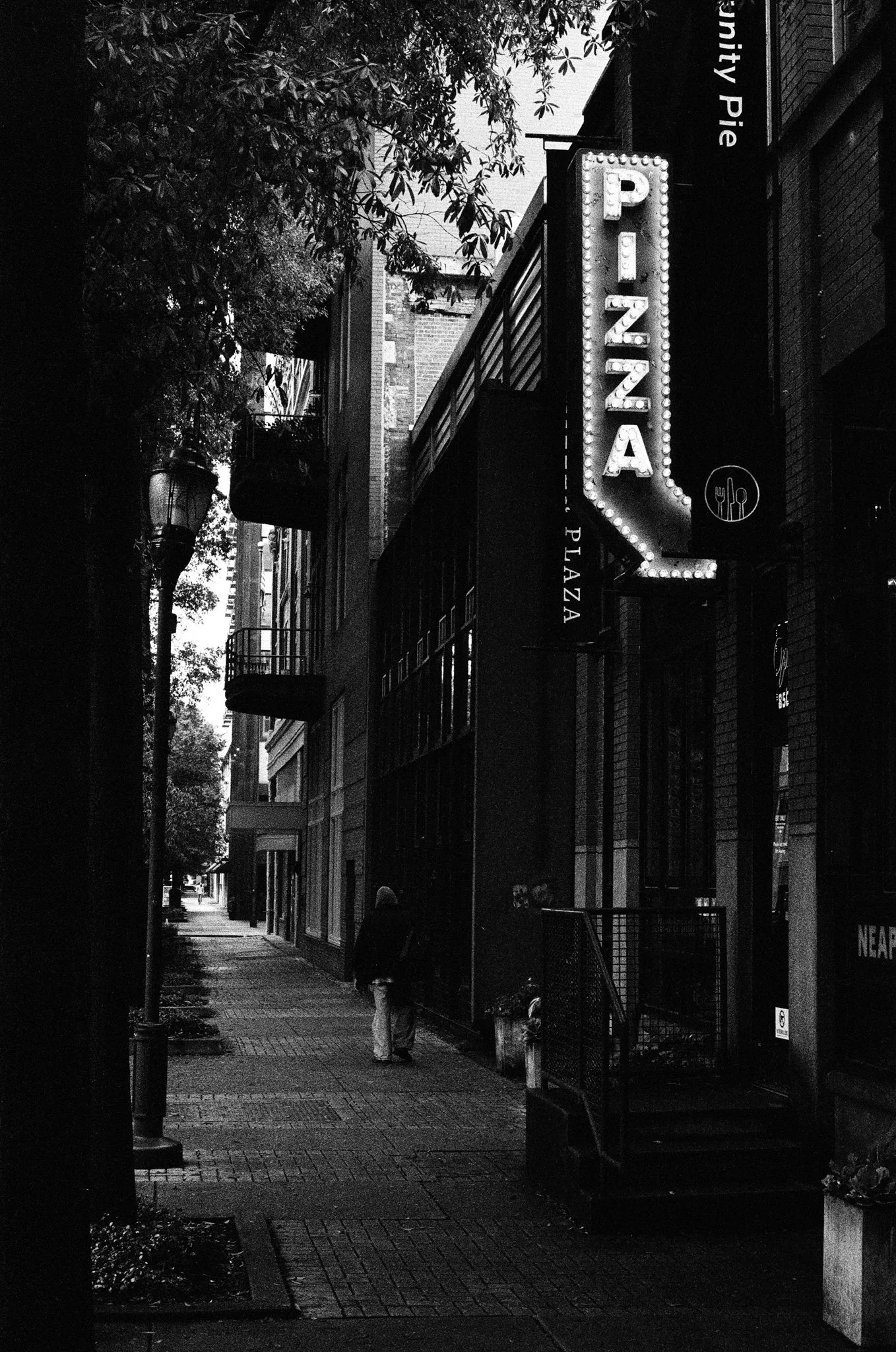 A black and white photo of an empty city sidewalk with a person walking away from the camera, lined with buildings, trees, and a parking sign.