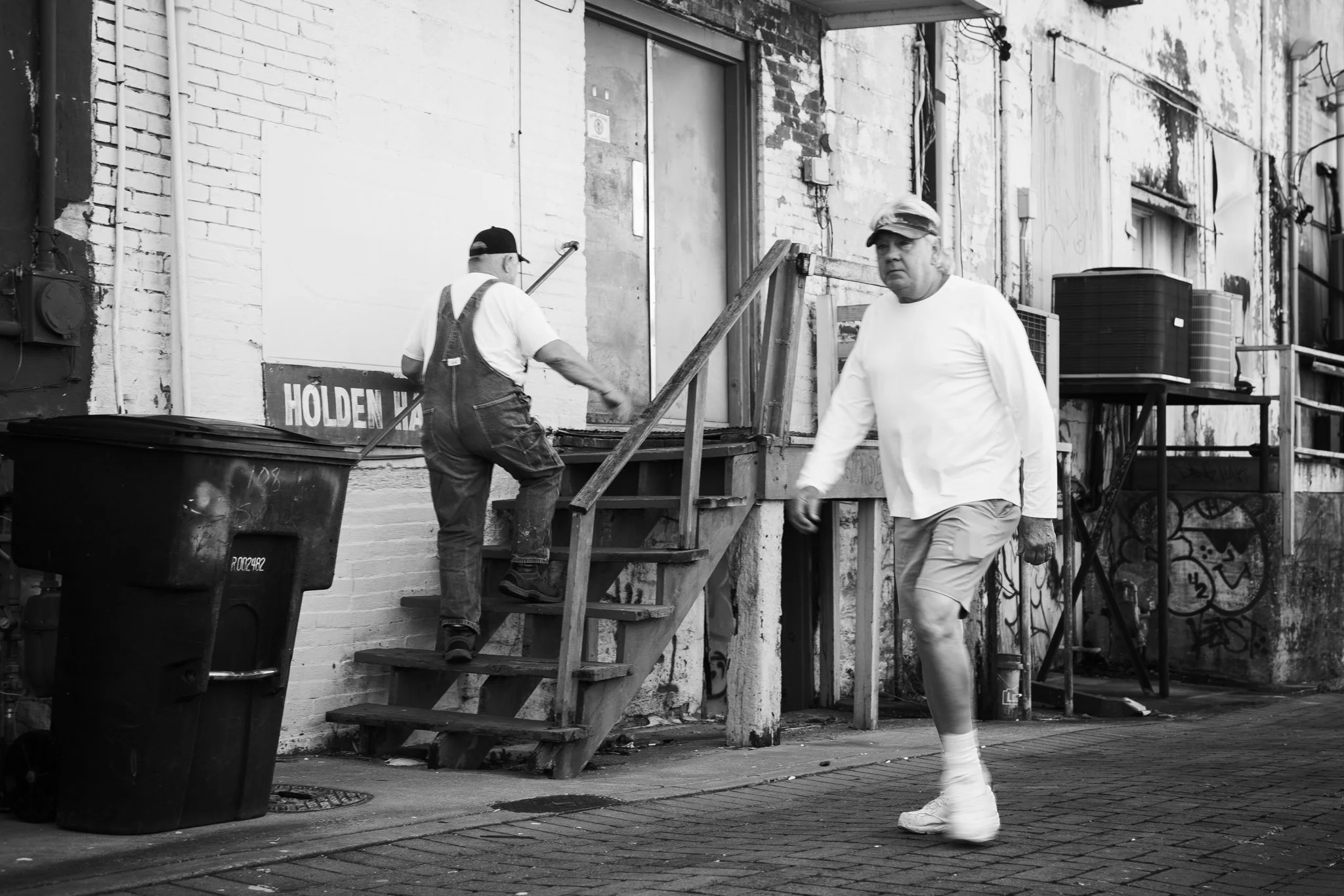Two men working outside a building with brick and concrete walls. One man is ascending stairs, wearing overalls and a cap, while the other man is walking past in a white shirt and shorts. There are large trash bins, an air conditioning unit, and graf