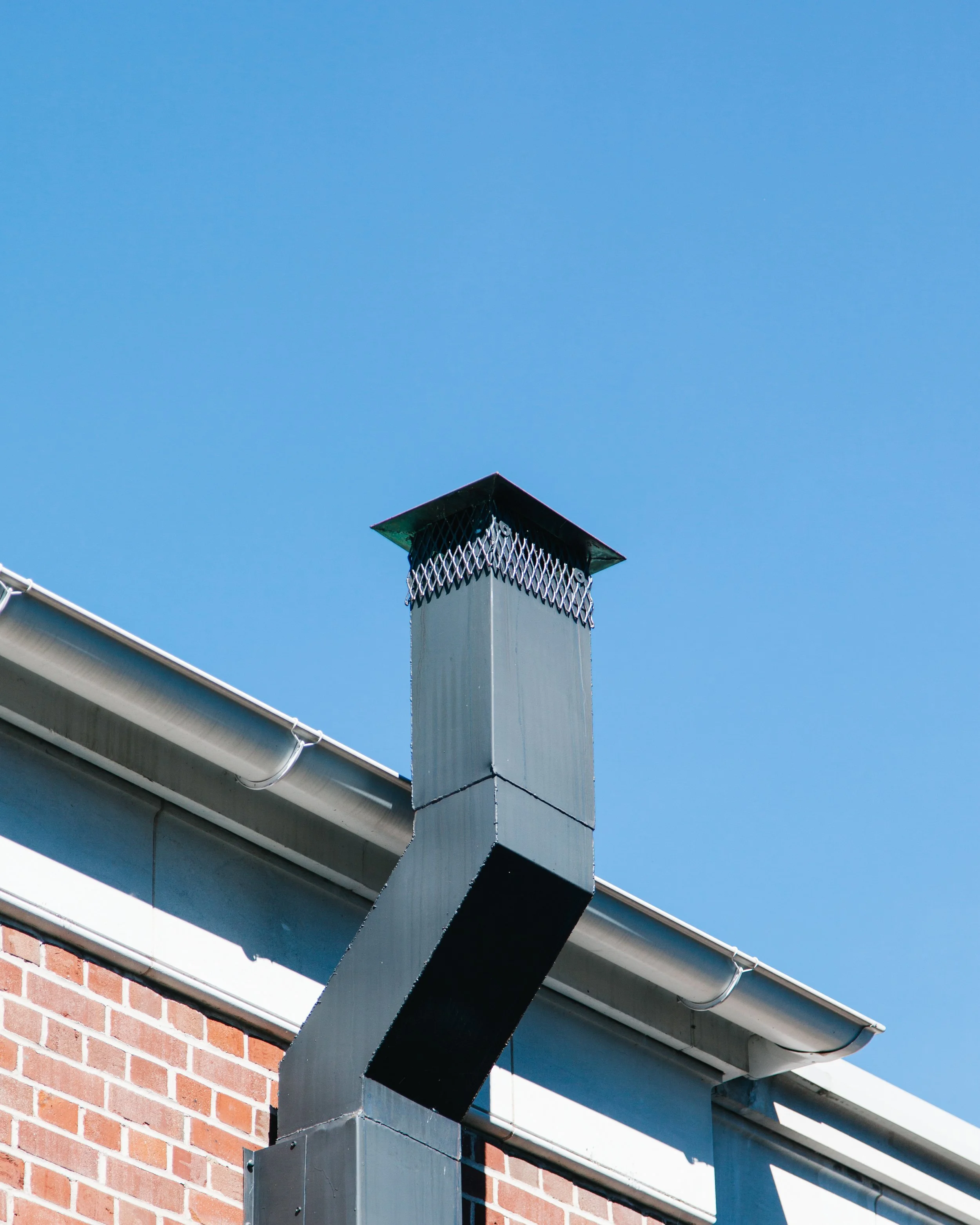 Close-up of a chimney flashing with a metal cap on a brick house, under a clear blue sky.
