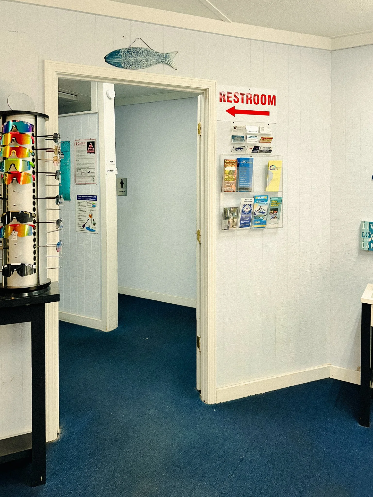 Interior of a building with a door leading to the men's and women's restrooms, a wall-mounted brochure rack, and a sign pointing to the restroom.