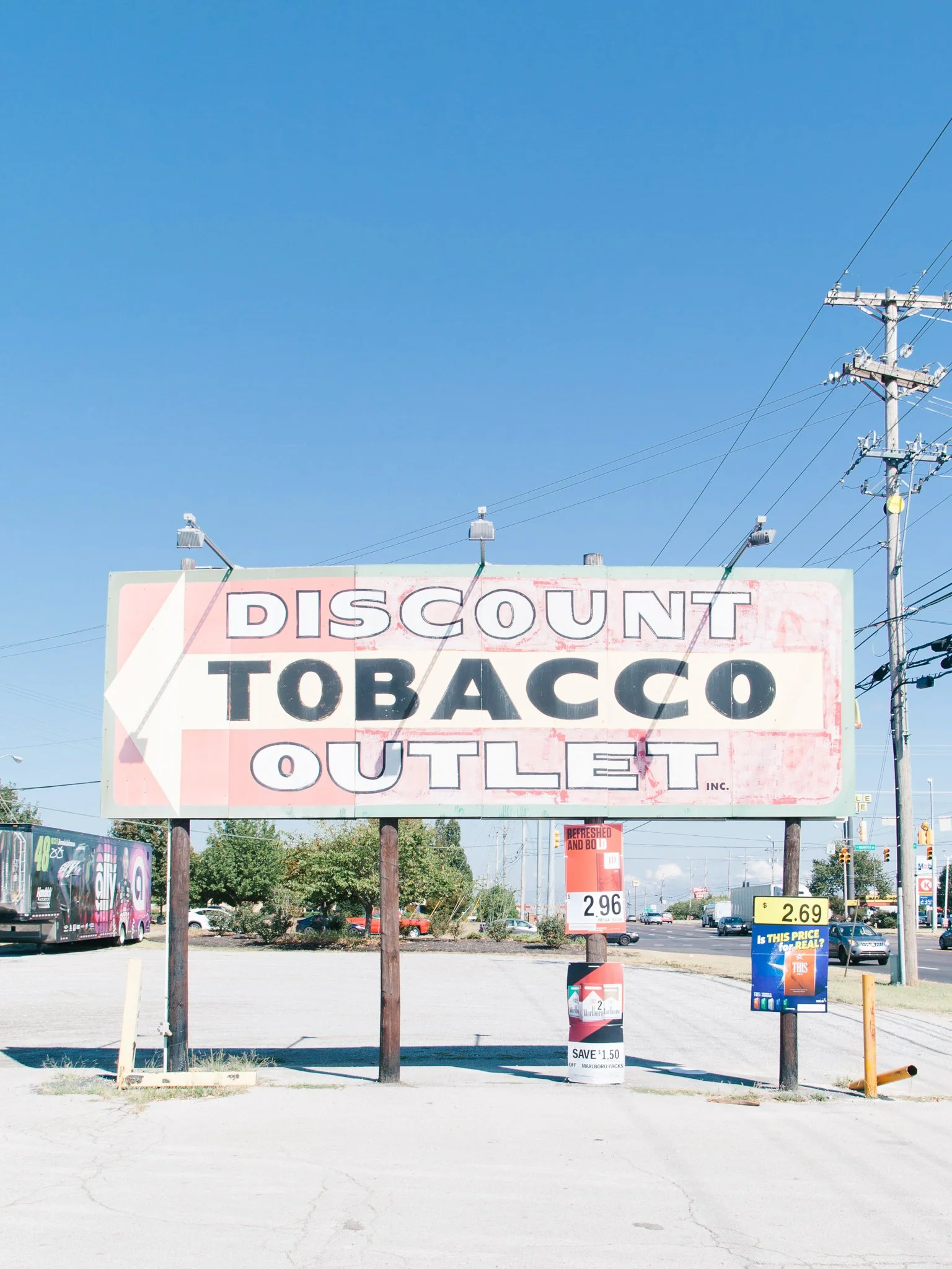 Large billboard advertising a tobacco outlet with the words 'DISCOUNT TOBACCO OUTLET' in bold letters, set in a parking lot under a clear blue sky, with power lines and some smaller signs and vehicles in the background.