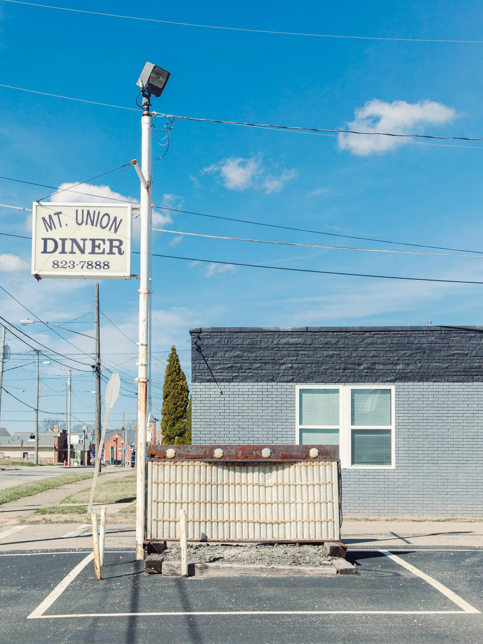A parking lot with a makeshift barrier, a gray brick building with white window frames, a tree, and a clear blue sky with a few clouds.