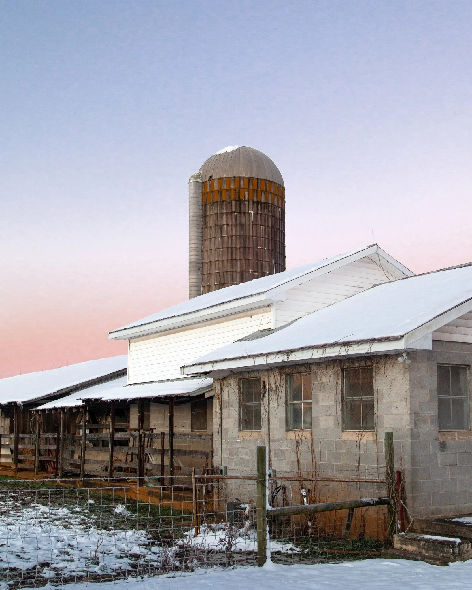 A house under construction with a chain-link fence in snowy weather. In the background, there is a barn or silo with a rounded top, rusted metal surface, and a small vent pipe. The house has white siding, large windows, and a sloped roof with snow on