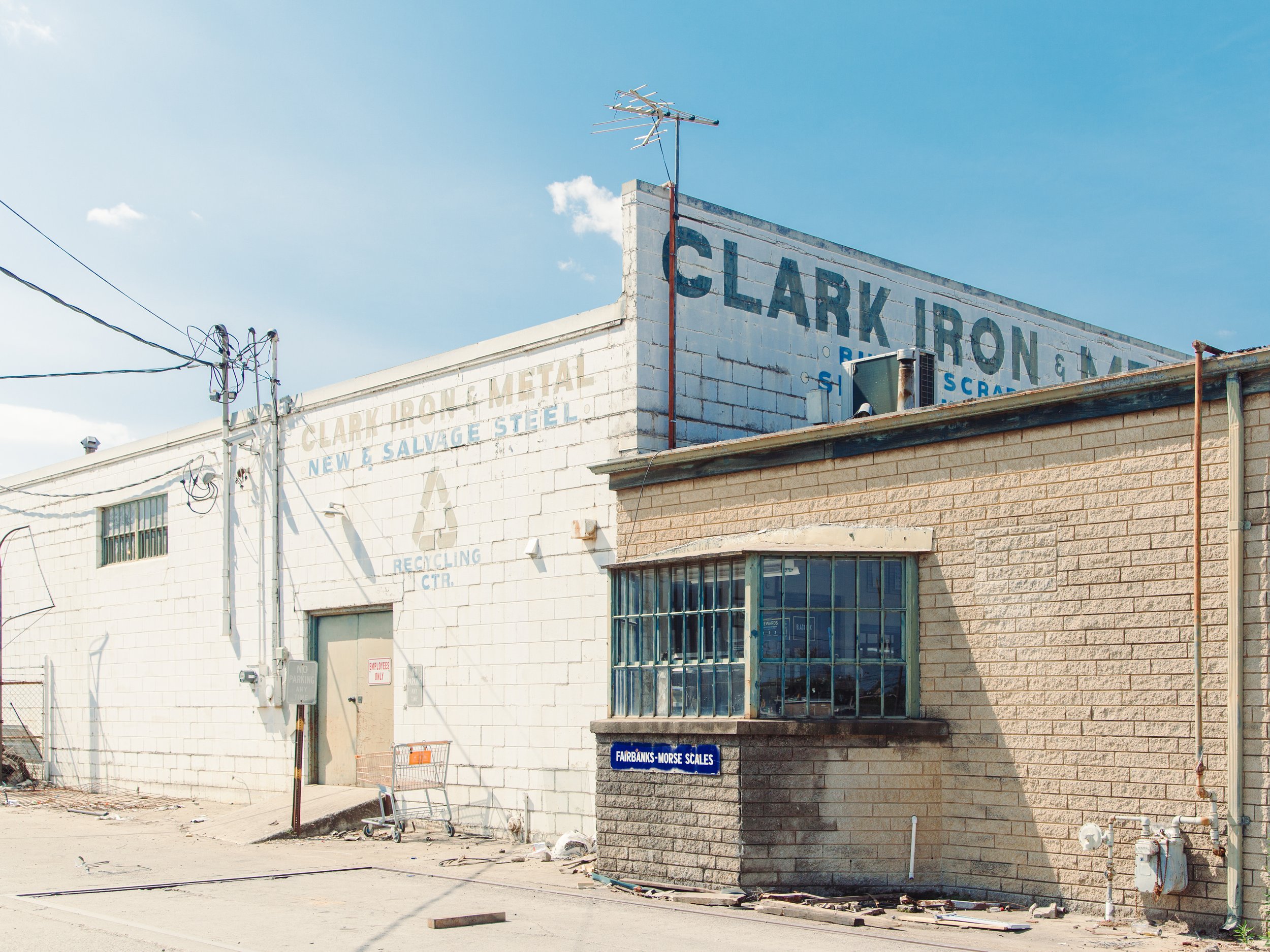 Side of a brick building with signs for a scrap metal and recycling business, including a blue sign for Fairbanks-Morse Scales, under a partly cloudy blue sky.