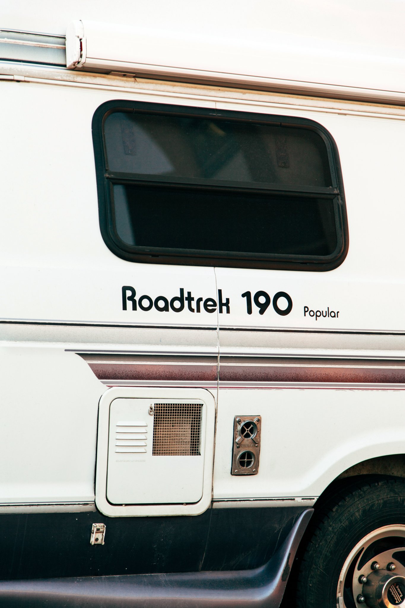 Close-up of the side of a recreational vehicle (RV) with a window and compartment door, brand name 'Roadtrek 190', and various vents and utility connections.