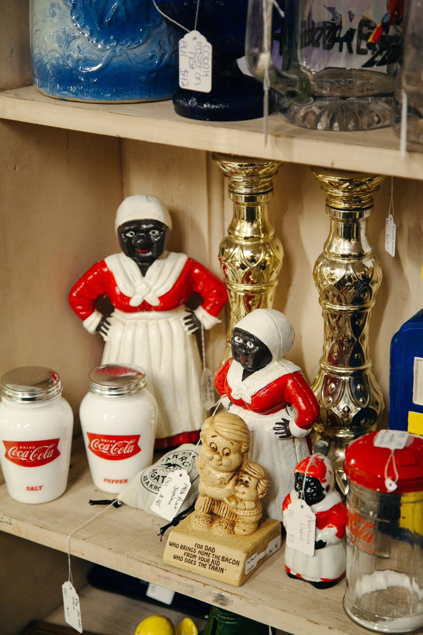 A wooden shelf displays decorative items including ceramics, vintage Coca-Cola bottles, and small figurines. Notably, there are two figurines dressed in red and white Santa-like costumes, with black faces and hands.
