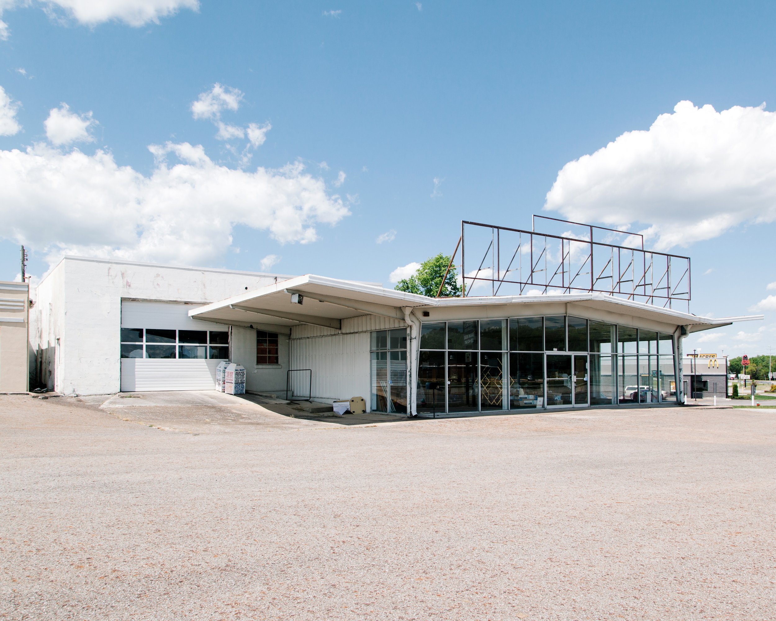 A building with a flat roof, large windows, and a trash bin outside, under a partly cloudy blue sky.
