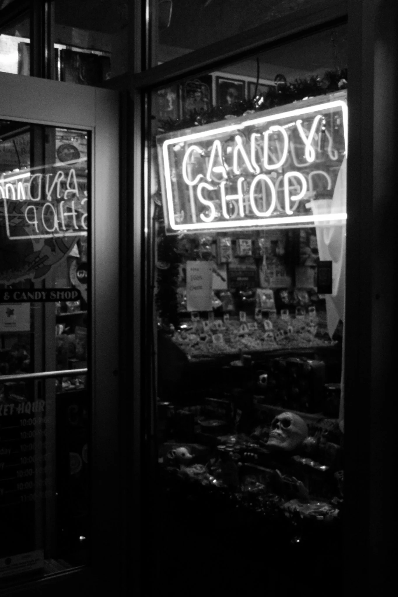A black and white photo of a candy and cookie shop at night, with a neon sign that reads 'Candy & Cookie Shop' in the window, and various candies and skulls inside.