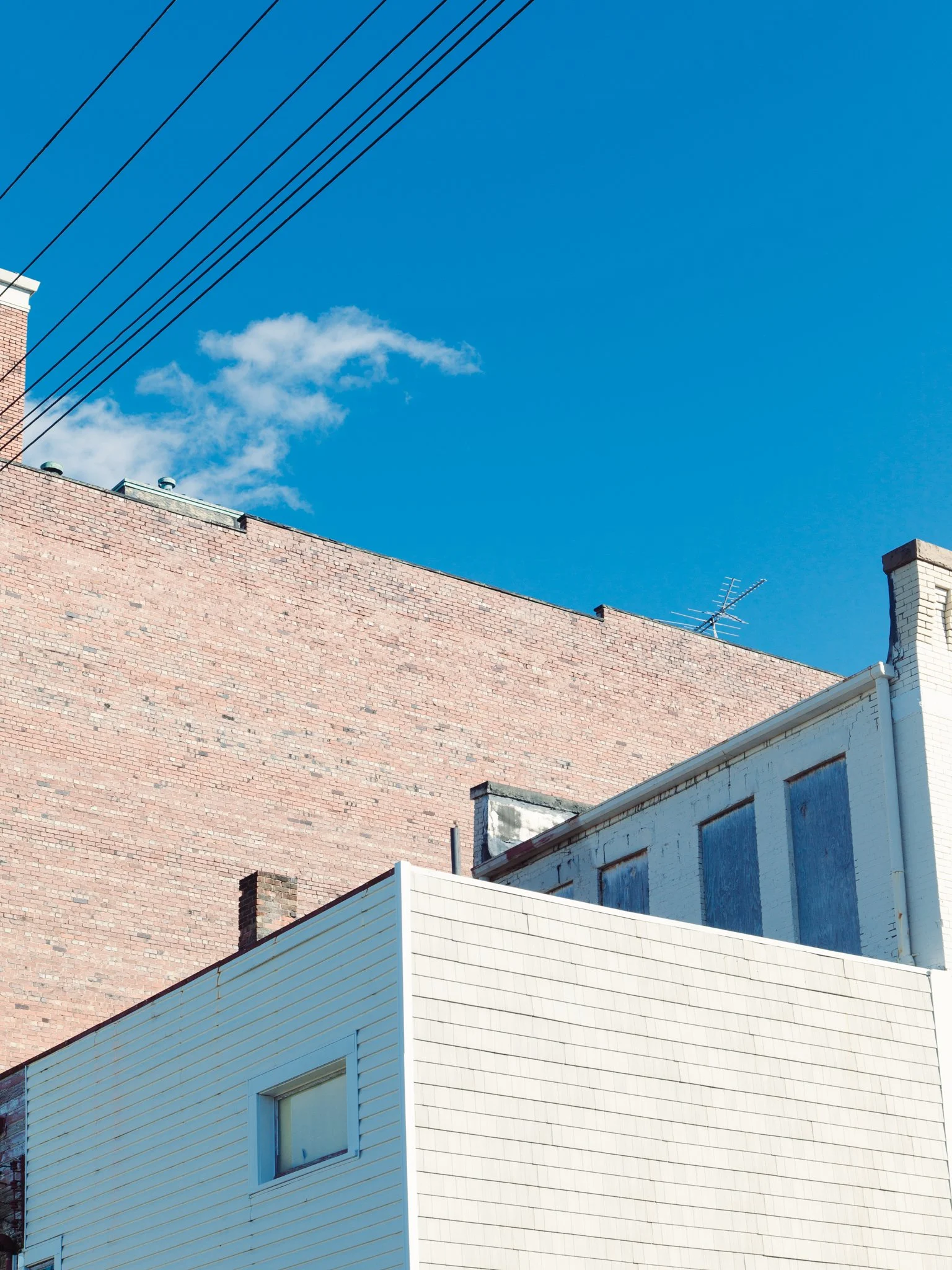 A clear blue sky with a few scattered white clouds, above rooftops of several buildings made of different materials including brick and siding, with visible antennas and vents.