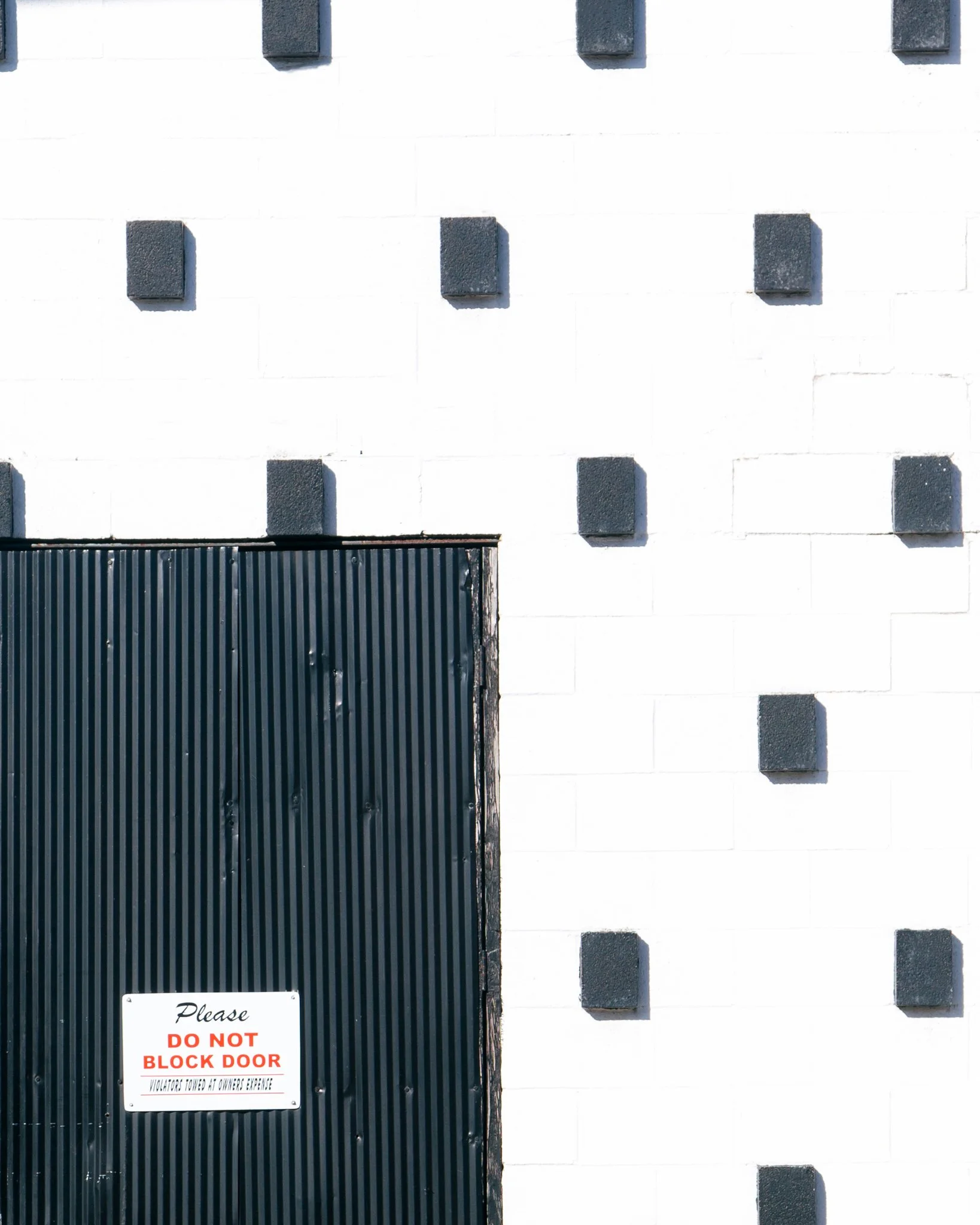 A white wall with a black metal door that has a section of vertical ridges. There are many dark gray rectangular blocks protruding from the wall in a regular pattern. A sign on the door reads: 'Please DO NOT BLOCK DOOR. Violators Towed At Owner's Exp