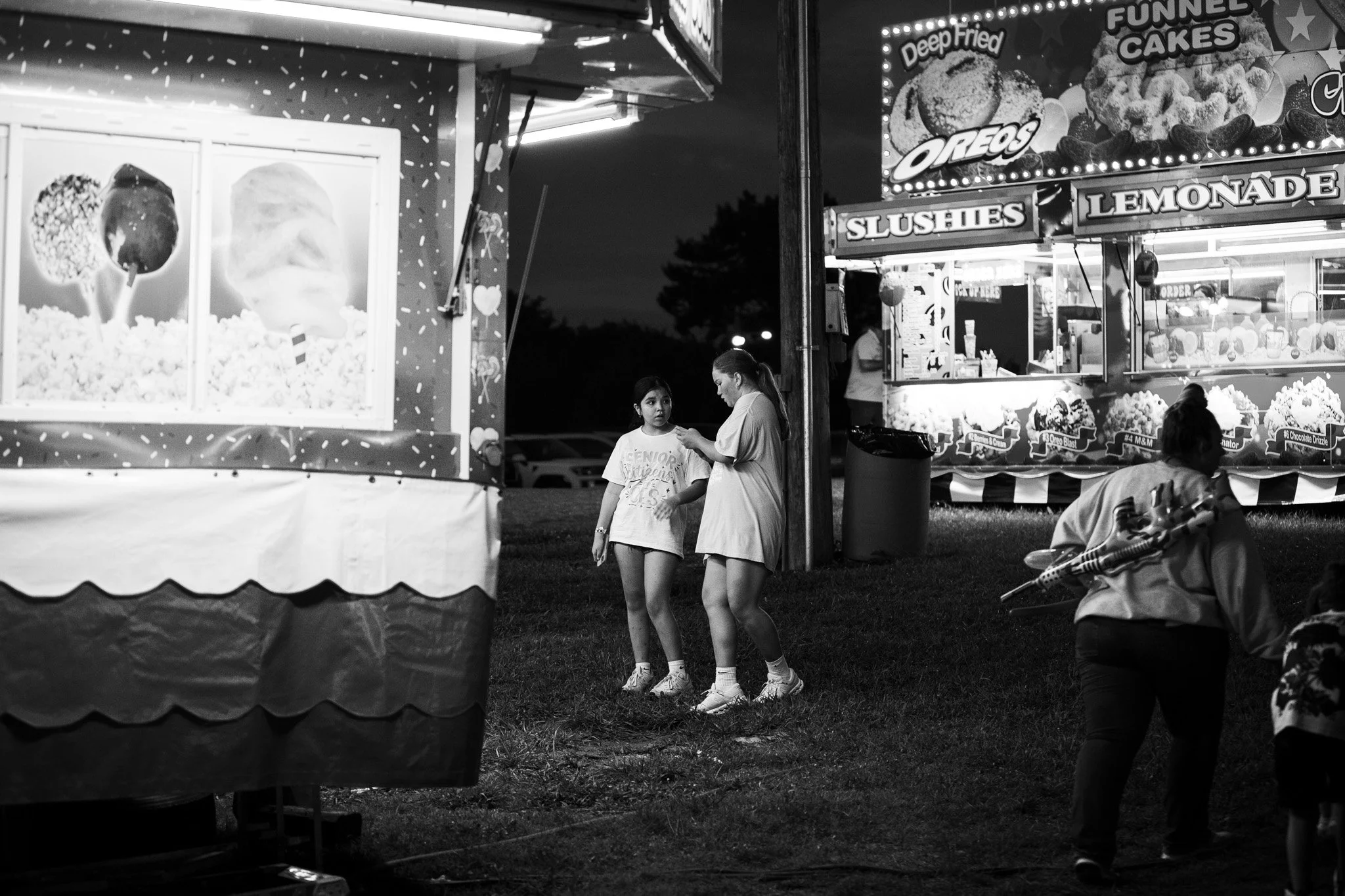 Night scene at a fair or carnival featuring food stalls with lights advertising deep-fried Oreos, funnel cakes, slushes, and lemonade. Two girls are standing and talking near a food stand, with other people sitting in the foreground and a dark sky wi
